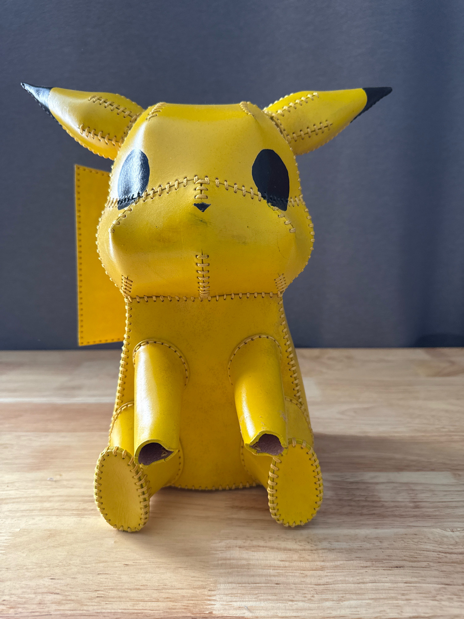 A leather, stitched Pikachu figure sits on a light brown wooden surface against a dark gray background. The figure is bright yellow with black accents on its ears and eyes. Its arms are bent, and its legs are tucked under its body. The figure appears to be handmade, with visible stitching along its seams. The lighting is soft, and the focus is on the Pikachu figure. The scene is devoid of any people or specific identifiable New York City landmarks or locations.