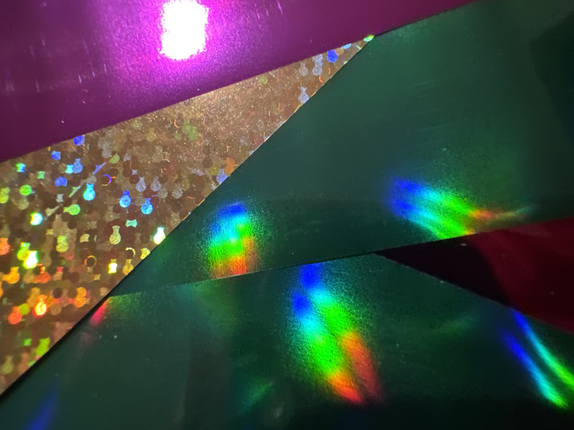 The image displays an abstract close-up of iridescent holographic materials. Three distinct layers are visible: a dark green material on the left, a shimmering bronze material with a textured pattern in the center, and a vibrant purple material on the right. Light reflects off these surfaces, creating rainbow-like patterns and bright highlights, particularly a prominent white glare on the purple material. The overall impression is one of shifting colors and light play, suggesting a creative or decorative context. No people, animals, or specific environmental features of Waterford, Ireland, are discernible.