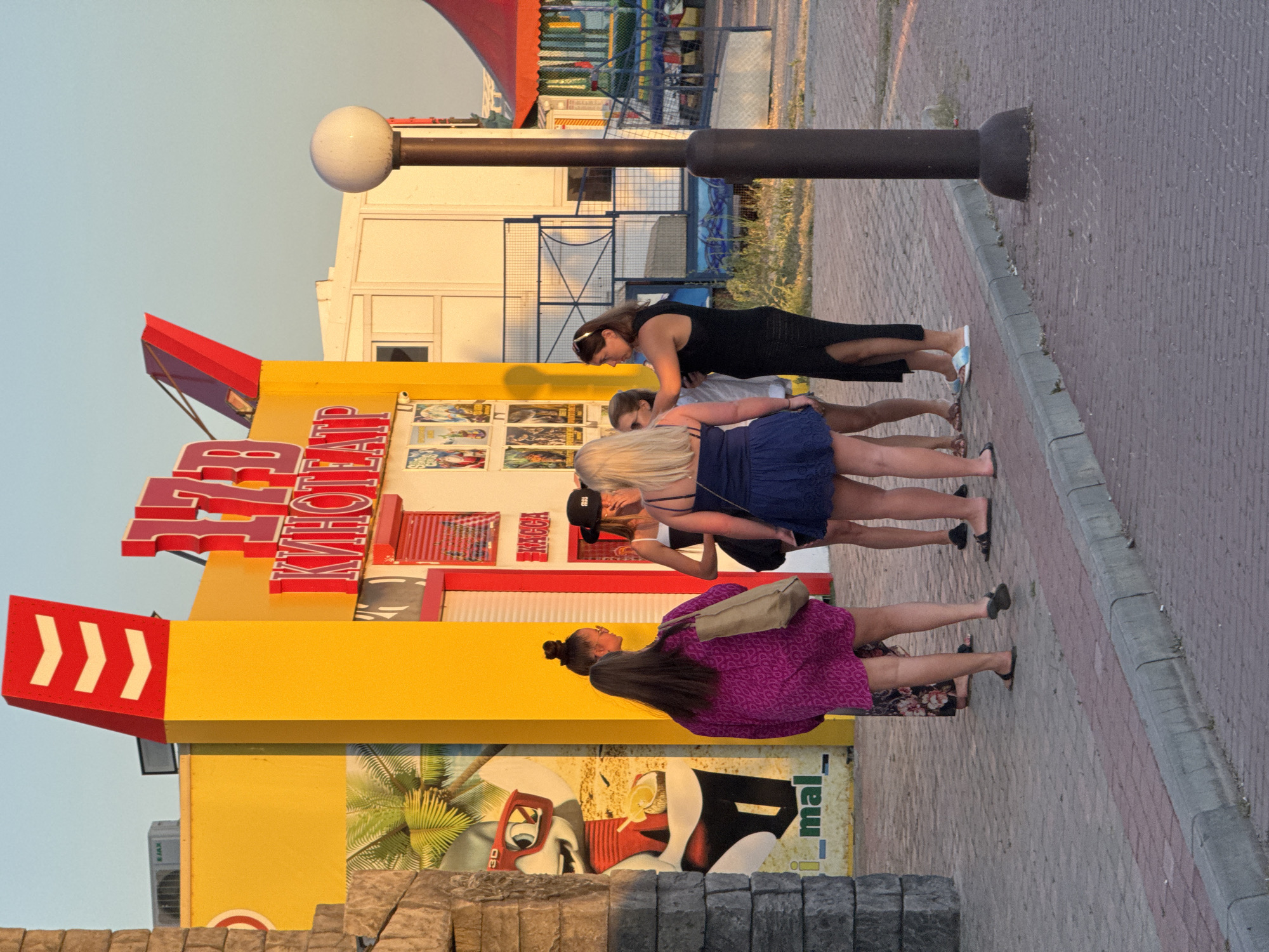 At the entrance to a small cinema named "17B KINOTEATR" in Novofedorovka, Ukraine, a group of five young women are gathered. The cinema building is painted bright yellow with red accents and features a large graphic of a cartoon character wearing 3D glasses. The women are dressed in casual summer attire, including dresses and shorts. Two of the women are wearing sunglasses. One woman on the right is wearing a black dress and the woman to her left is wearing a blue dress. The other three women are on the left side of the group. The scene is set on a paved sidewalk in front of the cinema, with a lamppost visible to the right. The sky is clear and blue, indicating it is daytime.