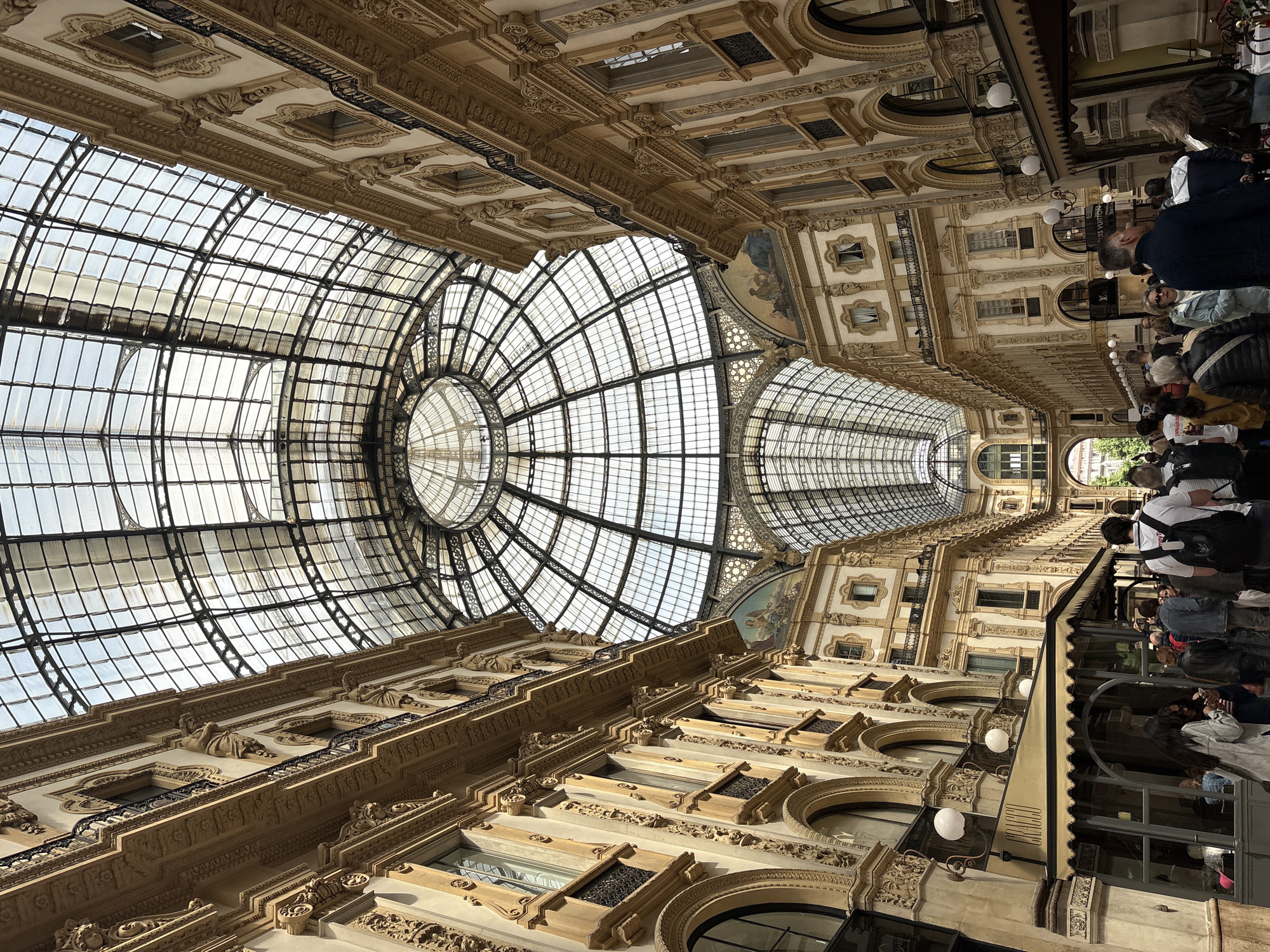 The image shows the interior of the Galleria Vittorio Emanuele II in Milan, Italy. The view is directed upward, focusing on the elaborate glass dome and the ornate architectural details of the surrounding buildings. The glass dome is structured in a radial pattern, allowing light to filter into the galleria. The buildings are adorned with intricate carvings and statues, showcasing a classic architectural style. People can be seen walking along the floor level of the galleria. The scene is bathed in natural light.