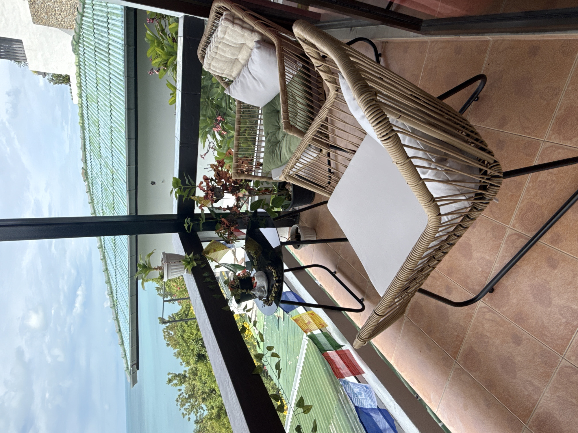 The image depicts a balcony overlooking a tropical seascape in Ko Samui District, Thailand. Two rattan chairs with cushions and a small black table are arranged on the tiled balcony floor. The table has a decorative white cat figurine and a small potted plant on it. Prayer flags in various colors are strung along the balcony railing, adding a touch of local culture. Behind the balcony, the green tiled roof of a building is visible, and beyond that, lush green trees and the turquoise ocean under a cloudy sky. The scene conveys a sense of relaxation and tropical ambiance.