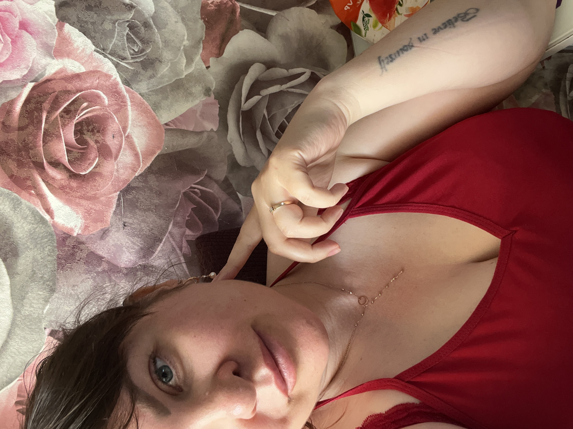 A woman with brown hair and blue eyes is taking a selfie. She is wearing a red top and a necklace. Her left arm is resting on her chest, and she has a tattoo on her right forearm that reads "Believe in yourself". The background is a floral wallpaper. The image does not contain any information that would allow for the determination of its location in Noviy, Russia.