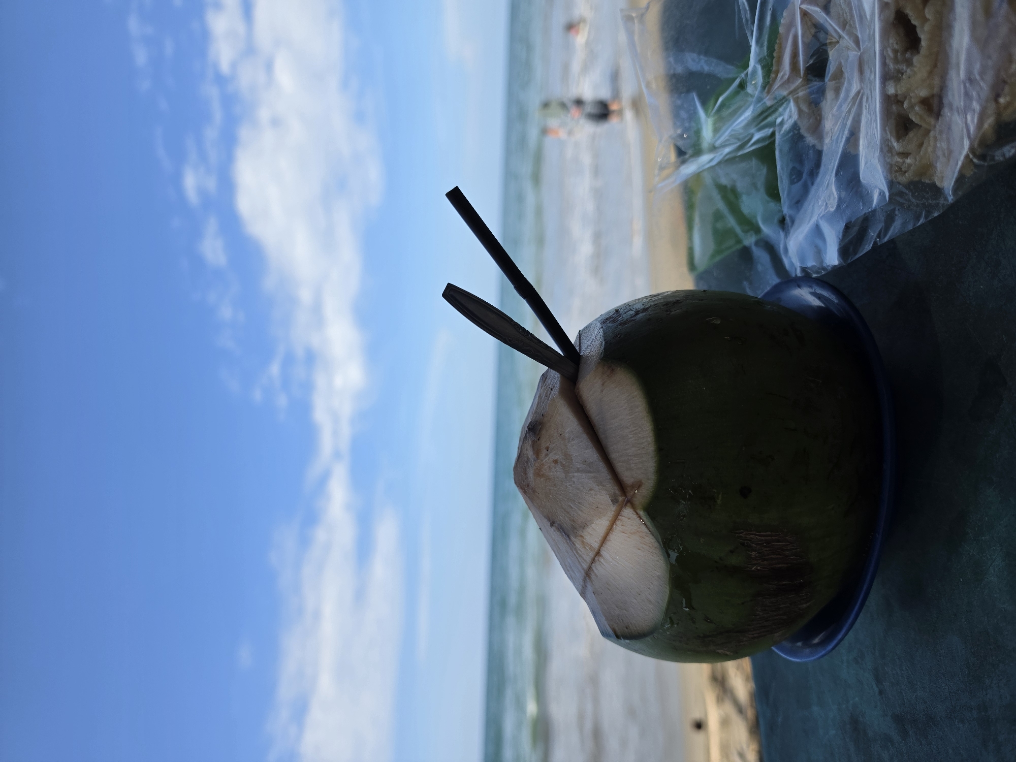 A young green coconut with two straws inserted is placed on a blue saucer on a dark blue surface. The coconut is positioned to the left of the frame, with its top cut open. In the background, the ocean meets the sky under a partly cloudy blue sky. Gentle waves are visible, and two figures are in the shallow water. To the right of the coconut, a clear plastic bag contains some light-colored snacks. The scene appears to be a beachside setting in Lhokseumawe, Indonesia, suggesting a relaxed atmosphere.