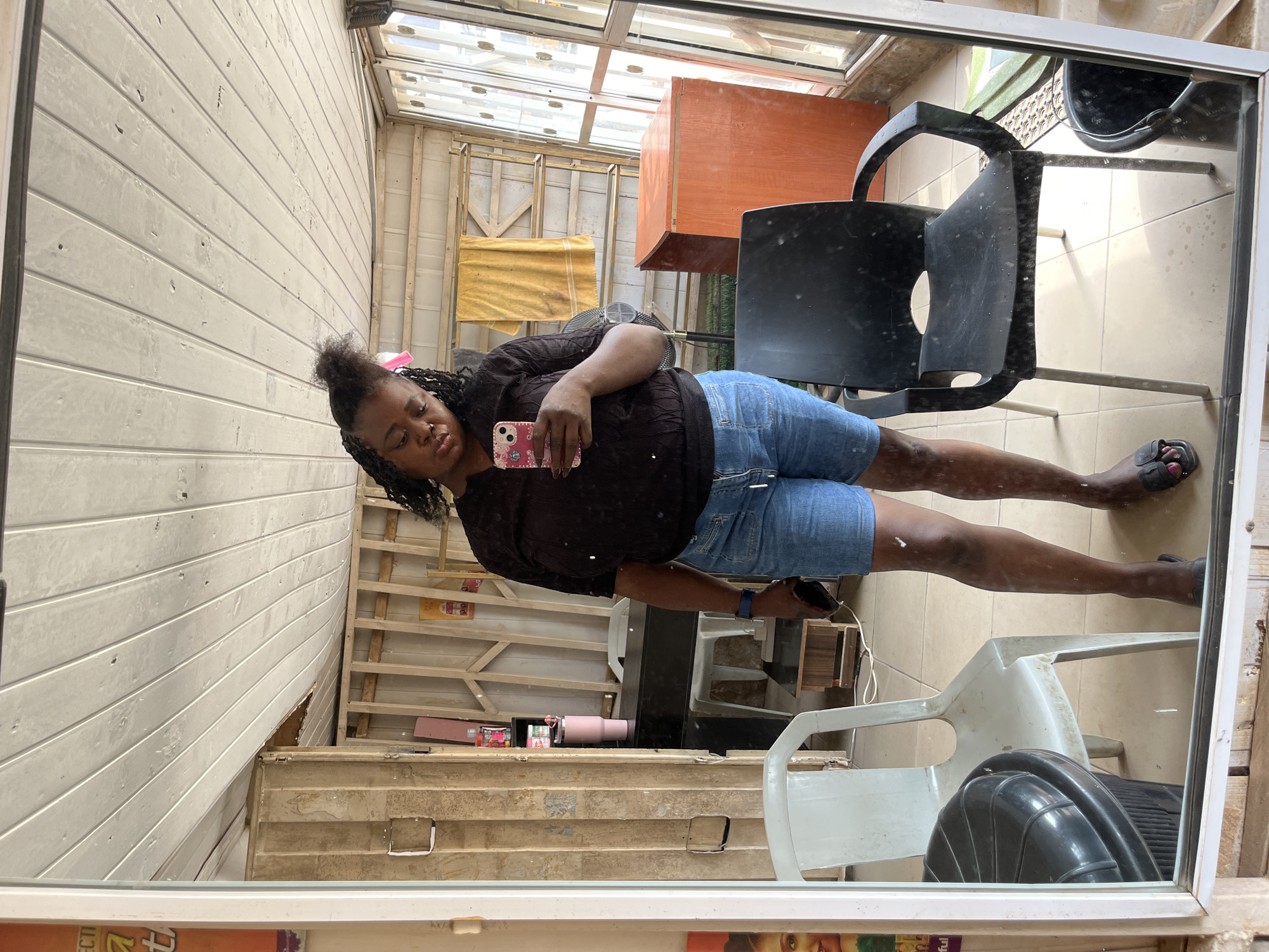 A woman is taking a mirror selfie. She has dark skin and dark curly hair styled in braids that are partially pulled up. She is wearing a dark brown, textured long-sleeved shirt and light blue denim shorts. She is holding a smartphone in her right hand, which is positioned in front of her, angled towards the mirror.
The setting appears to be an indoor space with a wooden plank wall. There is a large mirror reflecting the woman and the surroundings. Behind her, visible in the reflection, are various objects including a black chair, an orange cabinet, and a yellow piece of fabric. The ceiling is made of glass panels, suggesting a greenhouse or sunroom-like environment. The floor in the reflection is tiled. The overall impression is a casual, perhaps slightly cluttered, indoor space. There is no clear indication of the time of day or weather. The woman's expression is relaxed and neutral.
The scene is captured from a high angle, looking down at the woman and her reflection. The image is oriented in a way that makes the woman appear to be lying down or reclining, with her legs extending towards the bottom of the frame.
In the reflection, on a shelf-like structure in the background, there are several items including a pink tumbler and some bottles. Some text is visible on the bottom edge of the mirror frame and on items in the foreground of the reflection, but it is largely unreadable. However, the word "beautiful" can be partially seen on a magazine or book at the bottom of the frame.