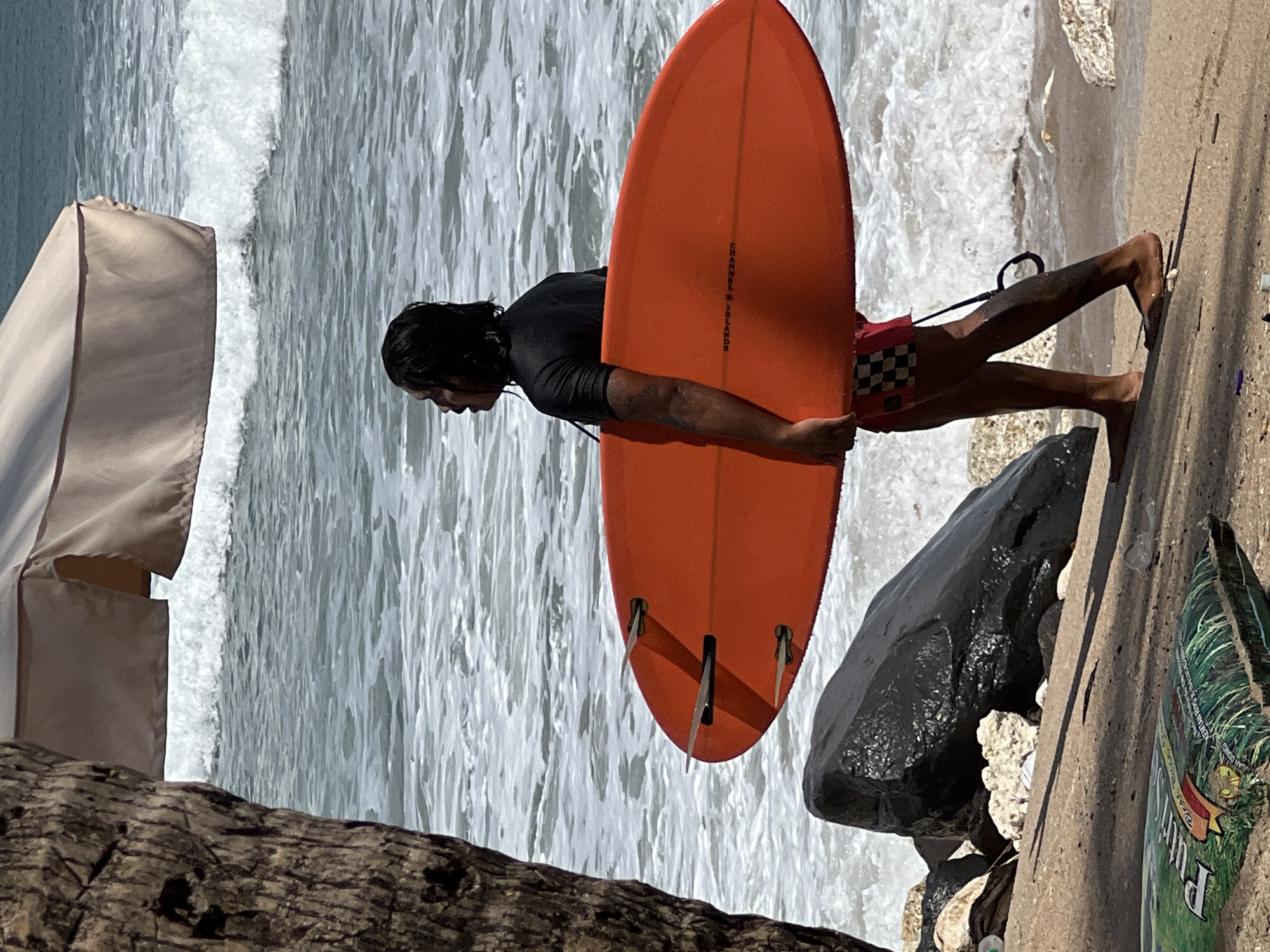 The image shows a person carrying an orange surfboard on a beach, likely in Kuta, Indonesia. The person is facing away from the camera, looking towards the ocean. They are wearing a black rash guard and red and white checkered swim trunks. The surfboard has three fins. In the background, waves are crashing on the shore. A large black rock is visible on the left side of the image, and a portion of a tree trunk is also present on the far left. A beach umbrella or canopy is partially visible at the top of the image. The sand on the beach appears damp, and there is some debris scattered on the ground, including a bag that reads "Putri" with text below it. The overall scene depicts a typical beach environment with a surfer ready to enter the water.