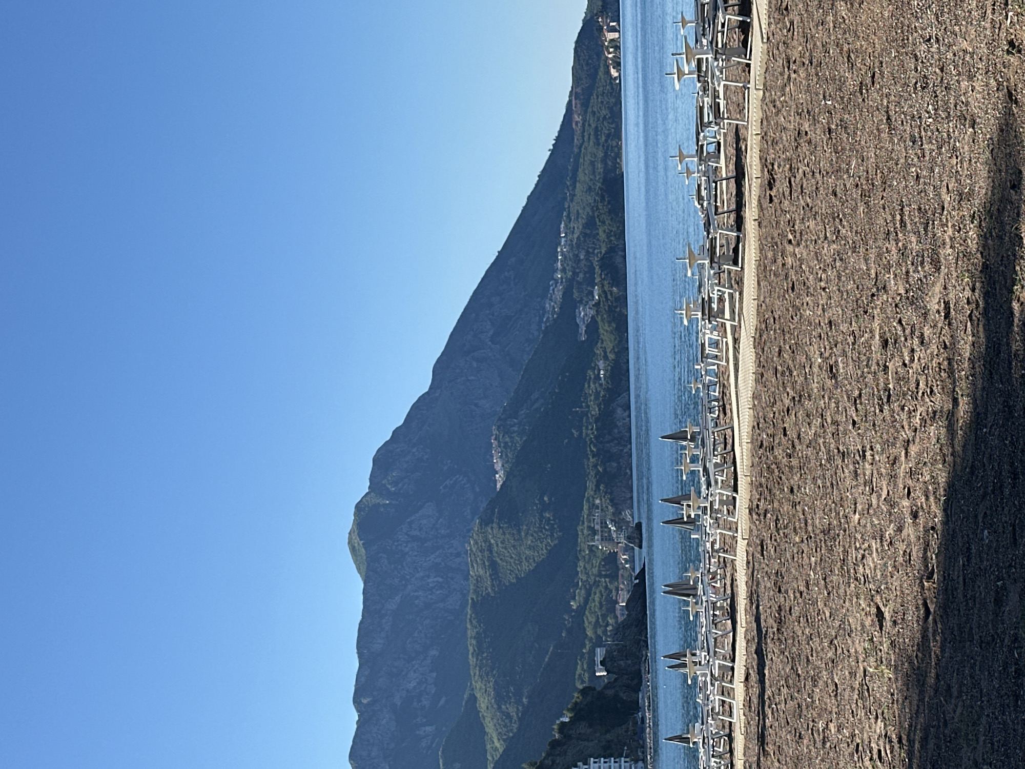 The image presents a landscape view from what appears to be a beach. A large mountain range is visible in the background, bordering the shoreline. The ocean extends from the beach towards the mountains. Numerous beach umbrellas and loungers are lined up along the shore. The foreground shows the beach's texture, indicating a coarse or pebbly surface. The overall scene evokes a coastal setting, possibly in the region of Boreti, Montenegro. No people or activities are clearly visible.