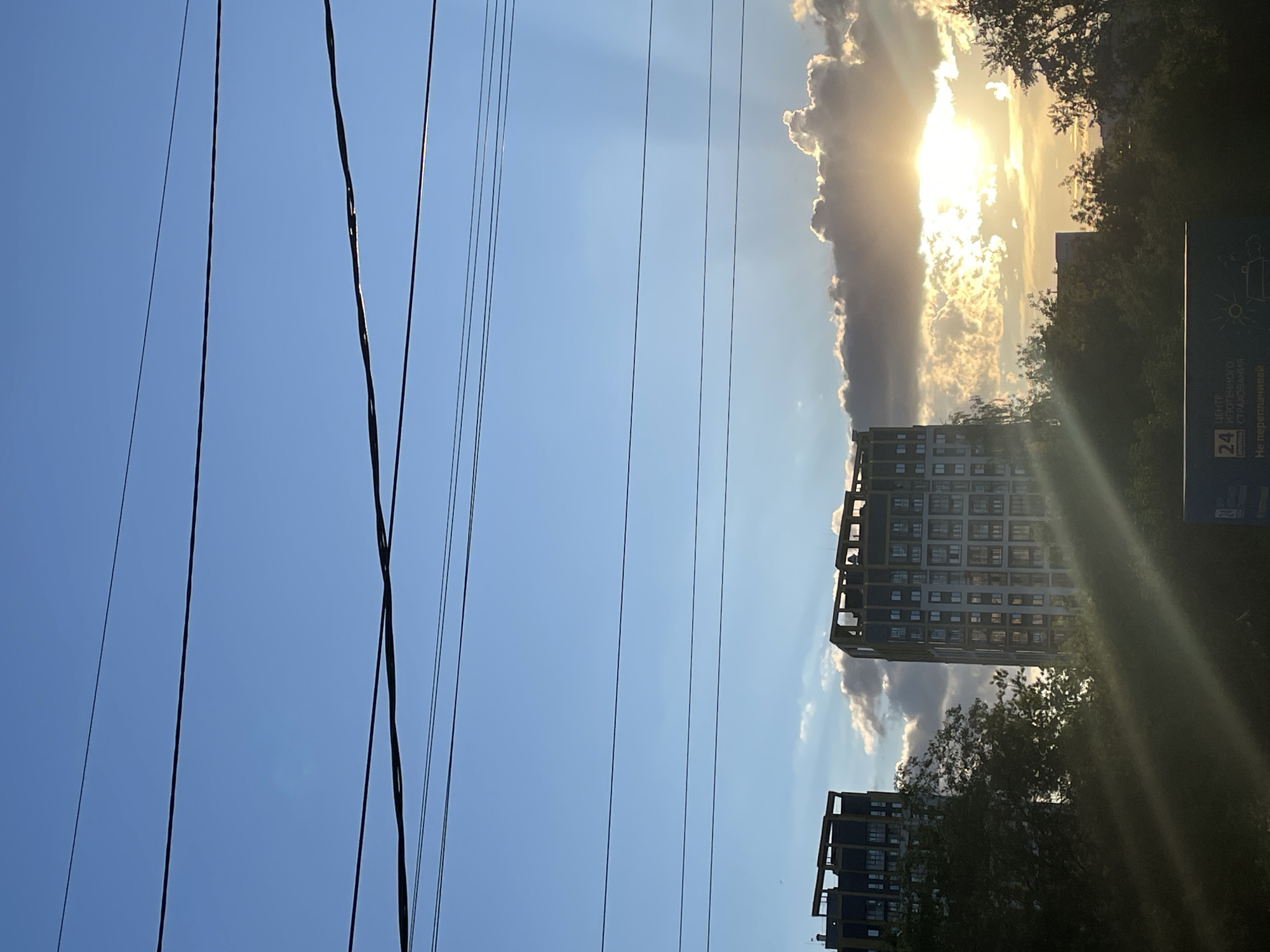 The image shows a view in Izhevsk, Russia, featuring power lines crossing a blue sky with clouds. Two high-rise buildings are visible, partially obscured by trees. The sun is shining brightly behind the clouds, creating a halo effect. A sign is present on the right edge of the image, with Cyrillic text and graphics.