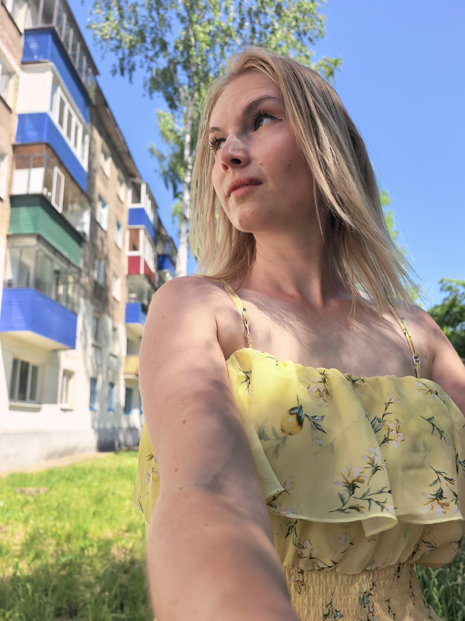 A young, blonde woman is the primary subject of this outdoor, medium-shot photograph, set against a backdrop identifiable as Sterlitamak, Russia. She gazes off to the left side of the frame, capturing a moment of apparent introspection. Her attire consists of a yellow floral summer dress with thin straps.
The background features a typical residential apartment building in Sterlitamak, easily recognizable by its architecture and color scheme. A tree stands to the right, its leaves adding a touch of nature to the urban setting. The clear, blue sky suggests a sunny day, casting shadows and defining the light in the scene.
The environment includes the apartment building, a tree, the ground with grass, and the sky. The interaction is the woman posing for the photograph, and the action is her looking towards something outside the frame. There are no other people or objects clearly visible.