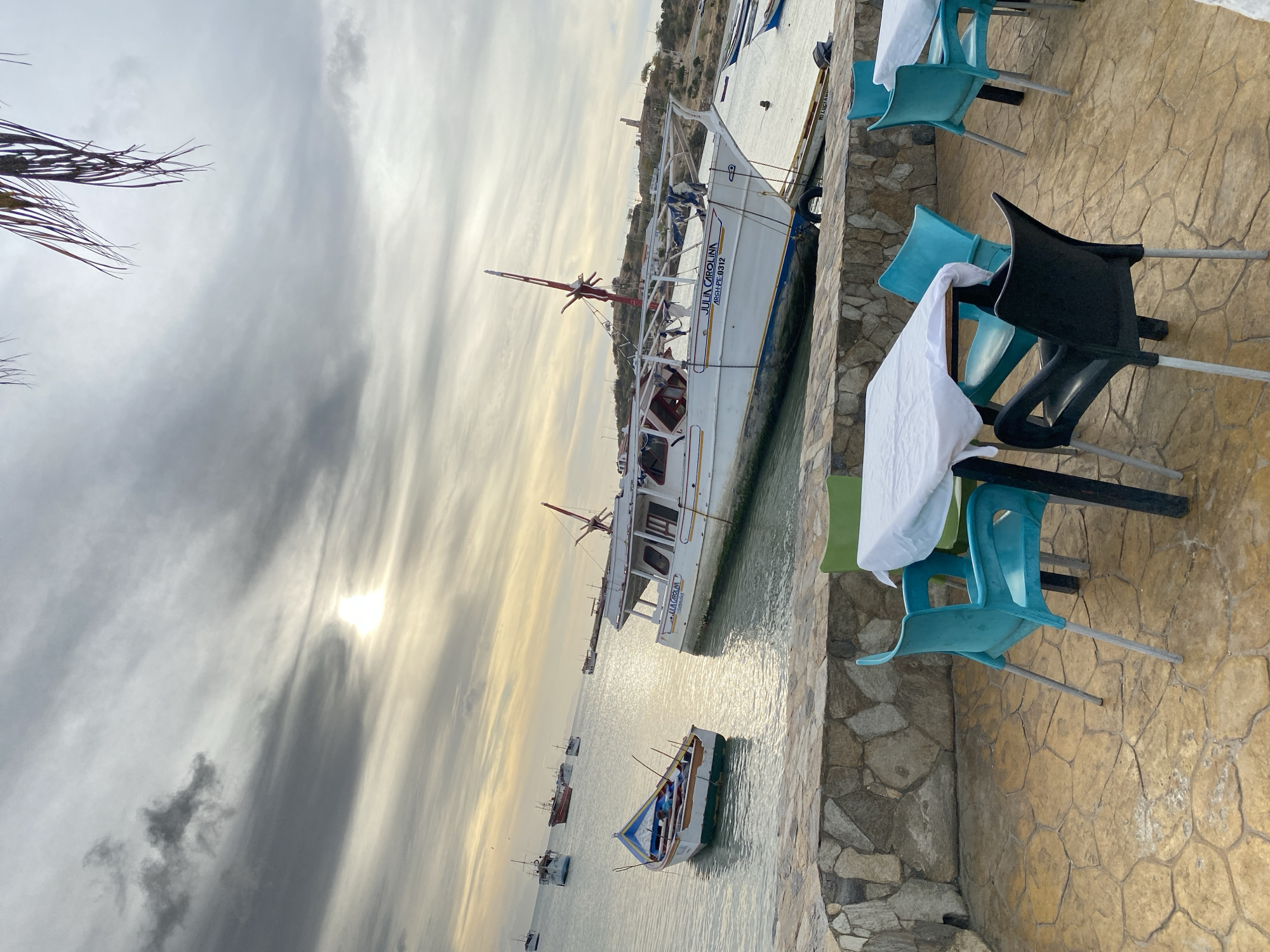 The scene depicts a coastal area in Juan Griego, Venezuela, viewed from a elevated patio. In the foreground, a paved patio floor, constructed from irregular yellowish-brown flagstones or stamped concrete, occupies the lower third of the image. A low, dry-stacked stone wall, composed of various light brown and grey rocks, separates the patio from the water. Two empty outdoor tables, covered with white tablecloths, are present. Surrounding the tables are five plastic chairs: three teal/turquoise, one black, and one green, all featuring metal legs.
The midground features calm water reflecting an overcast sky. A large white vessel named "JULIA CAROLINA" with registration "ARSH-PE-0312" is prominently positioned, partially grounded or moored in shallow water near the stone wall, listing slightly to its starboard side. It displays a red mast structure. To its left, a smaller, multi-colored wooden boat (white, blue, green, and red trim) floats, containing two seated figures. Several additional boats of varying sizes are anchored further out in the bay.
The background consists of a low, arid landmass or hill with sparse vegetation. A tall, slender, light-colored tower structure is visible on the distant landmass, along with what appears to be a road. The sky is largely overcast with multiple layers of grey and white clouds, and a diffused, bright patch of sunlight is visible towards the horizon on the right. Dried palm fronds hang into the frame from the top, indicating overhead foliage. No direct human action or interaction is evident beyond the two figures in the small boat.