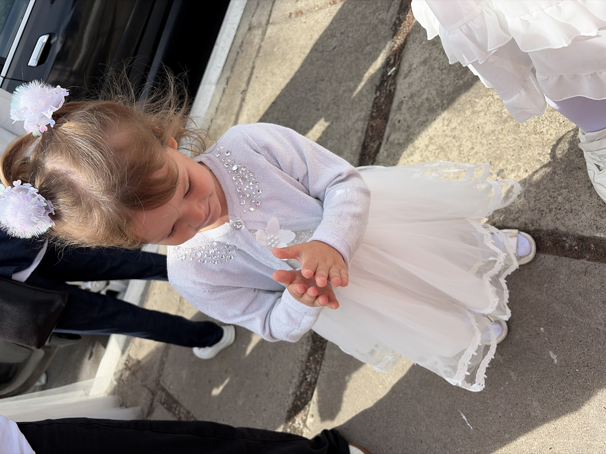 A young girl, wearing a light-colored dress adorned with glitter and a flower detail on the front, is standing outside. She has her hands clasped together, and her hair is styled in two pigtails with fluffy white clips. She is wearing white shoes. The background shows a paved surface and a portion of a black vehicle. The image does not provide enough information to determine a specific location within the city of Chaikovsky, Russia.