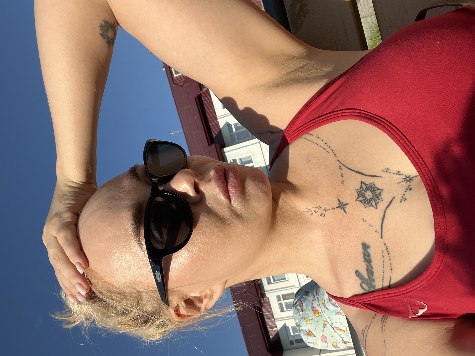 A medium shot captures a woman in a red top with sunglasses and her hand on her head. She has blonde hair pulled back in a bun. The woman has tattoos on her chest and arm. In the background, a building with a red roof and white walls is visible, along with a clear blue sky. A small child's hat with a unicorn pattern is on the left side of the frame. The image appears to have been taken outdoors in daylight.