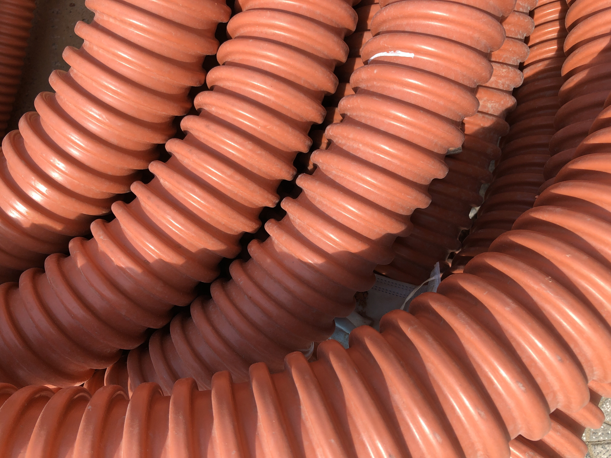 The image shows a close-up, overhead view of a pile of orange corrugated plastic pipes. The pipes are laid out in a coiled fashion, filling the frame. The texture of the pipes is emphasized by the lighting, which casts shadows within the corrugations. The environment appears to be outdoors, with a hint of a rough, possibly concrete surface visible at the bottom edge of the image. No people or specific landmarks are visible to indicate a precise location in Đà Nẵng, Vietnam.