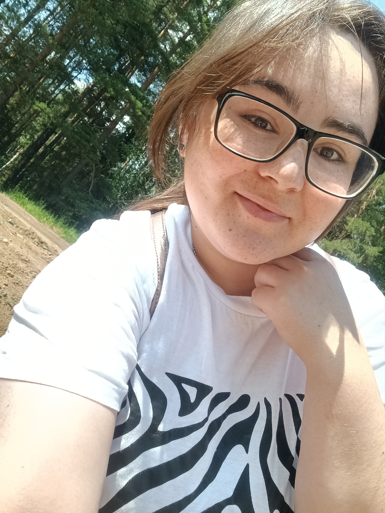 A young woman with freckles and glasses takes a selfie outdoors. She is wearing a white t-shirt with a black zebra stripe pattern. Her hair is brown and pulled back. She is resting her chin on her hand. The background shows a dirt path and a dense forest with tall evergreen trees, suggesting a rural or park setting outside of Noviy, Russia. The lighting indicates it is daytime.