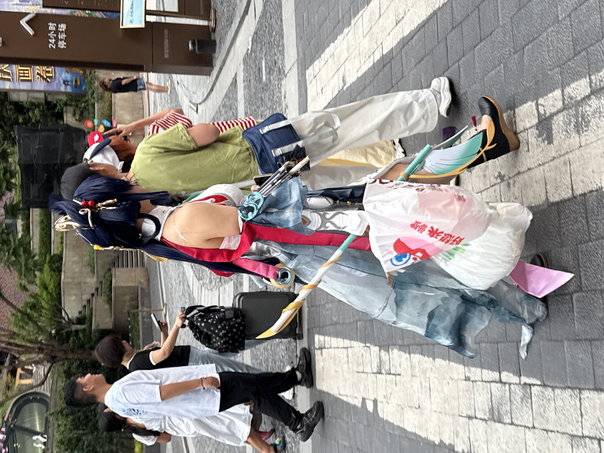 In Yuzhong District, China, pedestrians traverse a crosswalk. A person in the foreground is dressed in a detailed cosplay outfit, complete with an elaborate blue wig adorned with golden accessories, a white and red garment, and a flowing light blue skirt with a dark blue pattern. They carry a prop weapon resembling a sword and are holding a white plastic bag with pink writing. To their left, a man in a white t-shirt and black pants walks with another person in a black t-shirt and jeans. Behind them, a woman in a white dress walks alongside a black suitcase. Further back, a person in a green t-shirt and white pants walks with a striped shirt visible underneath. The environment features a paved sidewalk, street, and buildings in the background, including a sign with Chinese characters and "24小时停车场" (24-hour parking lot).