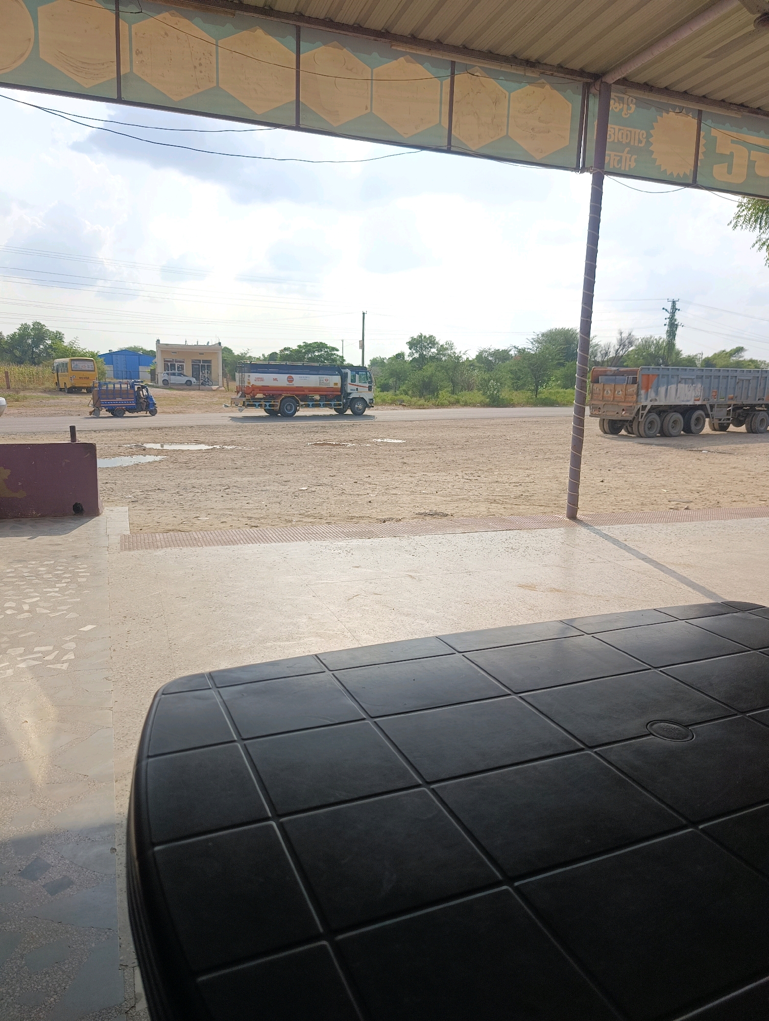The image shows a scene in Chhapri Bari, India, featuring a roadside view. In the foreground, a dark, textured table is partially visible. Beyond the table, a paved area leads to a sandy, dirt road. Several vehicles are present on the road. On the left, a yellow school bus and a blue auto-rickshaw are parked. Further down the road, a tanker truck with "ML" branding is driving, and a large, grey dump truck with multiple axles is parked on the right side of the road. The background consists of a line of green trees and vegetation, with a cloudy sky overhead. A building with a white facade and a smaller blue structure are visible behind the vehicles. A metal pole, wrapped with white and brown twine, stands near the dump truck. The scene appears to be an outdoor setting, possibly a rest stop or a commercial area along a highway.
