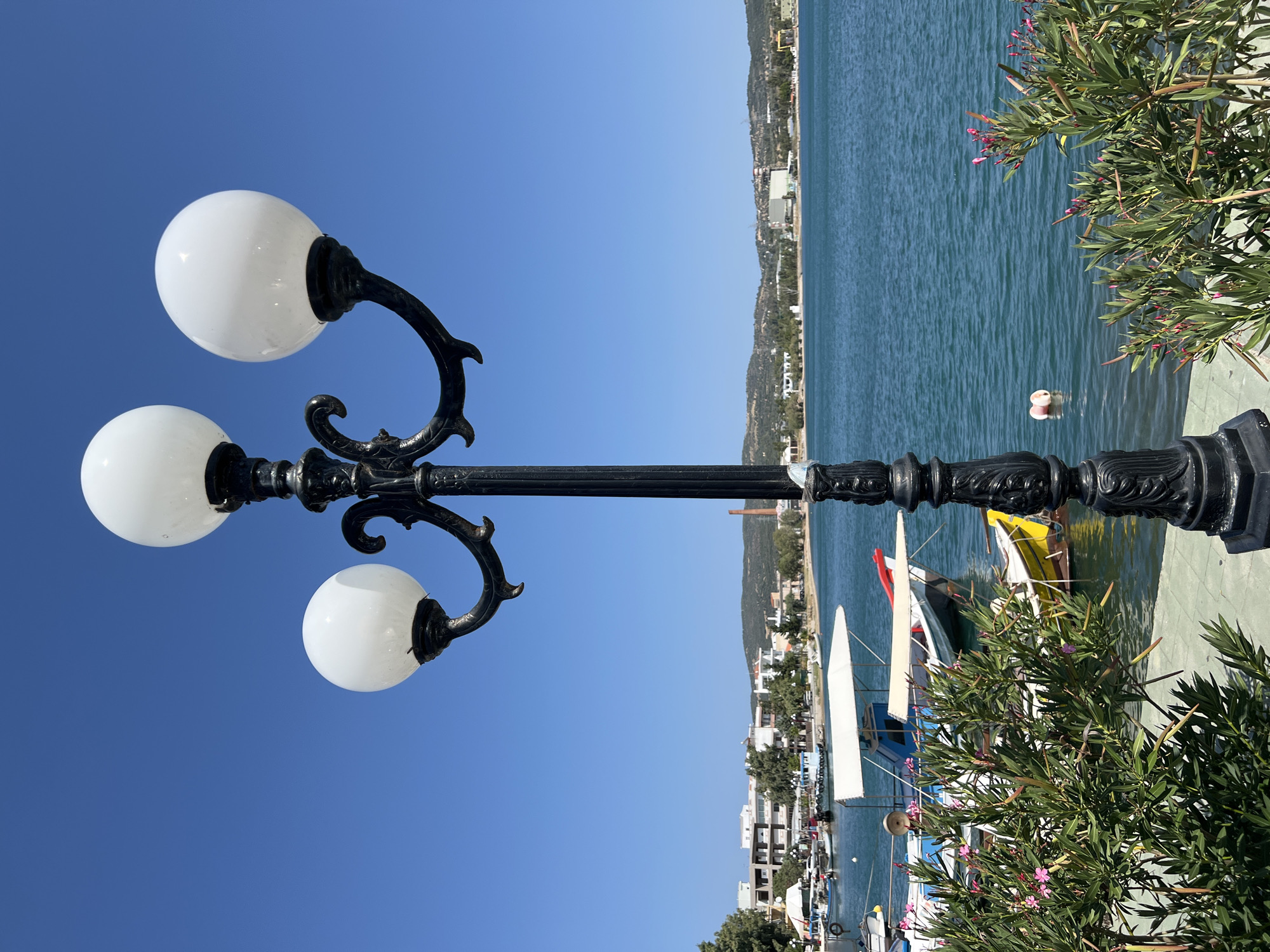 In Karavos, Greece, a black ornamental lamppost with three white globes stands in the foreground, casting a shadow on the green and grey tiled pavement. Behind the lamppost, a calm, blue body of water is visible, with several small boats docked. Some of these boats have white canopies providing shade. Lush green bushes with small pink flowers are also present along the water's edge. Across the water, a shoreline is lined with buildings of various sizes and architectural styles, interspersed with trees. In the distance, rolling hills form the horizon, with a clear, bright blue sky overhead.
