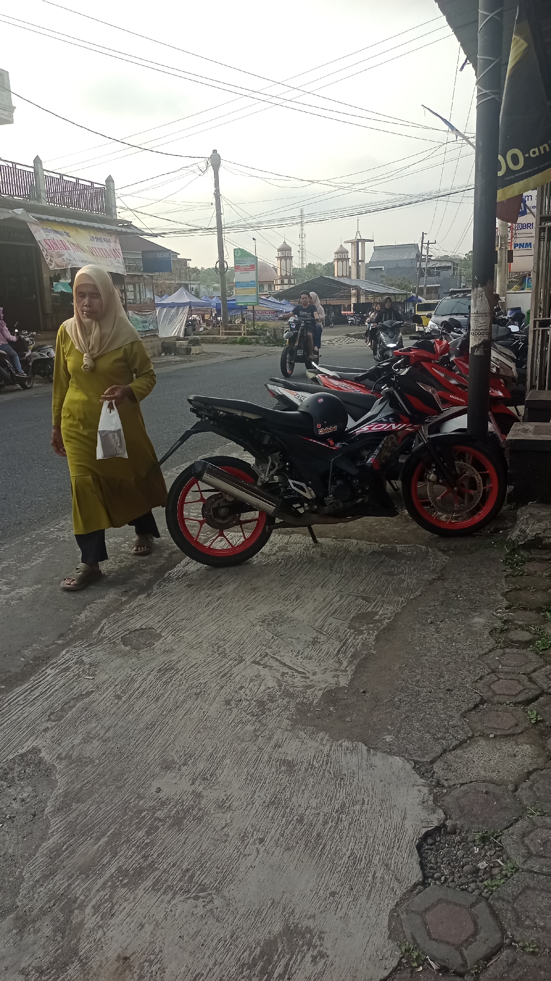 A woman in a yellow hijab and long dress walks on a concrete sidewalk next to a street in Karyamandala, Indonesia. She carries a white plastic bag in her right hand. Several motorcycles are parked along the street, with a black and red motorcycle prominently featured in the foreground. In the background, buildings with domes and towers suggest a religious or cultural presence, possibly a mosque. The sky is overcast, and power lines crisscross overhead. The scene depicts a typical street view in a town, with elements of daily life and urban infrastructure.