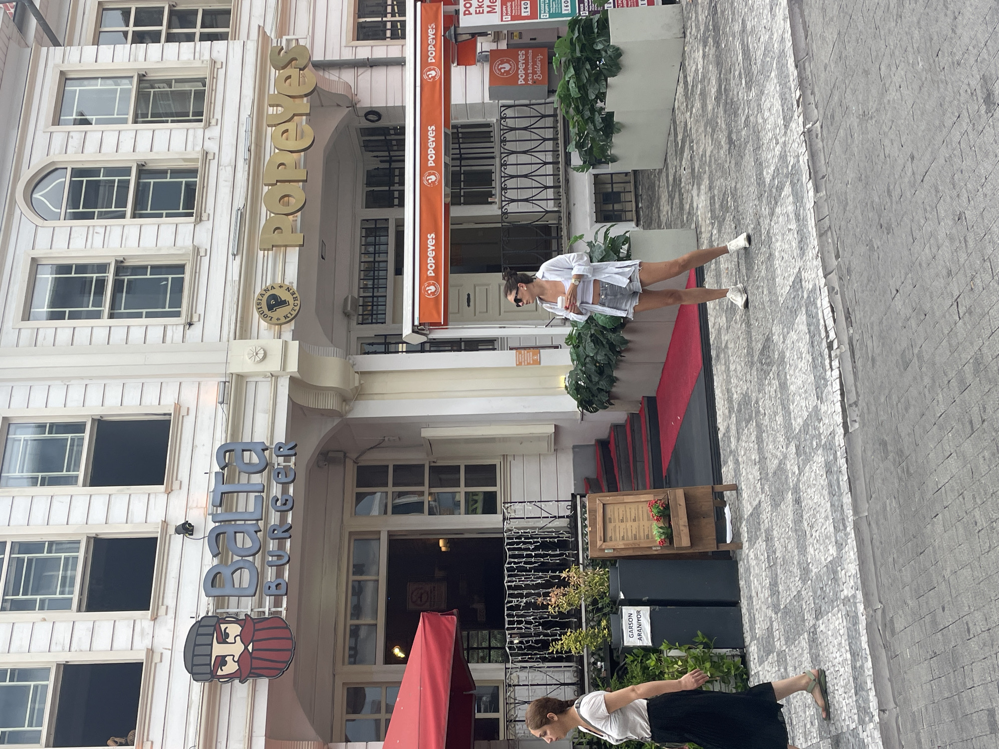 The image captures a street scene in Kadıköy, Turkey, featuring two prominent food establishments: Balta Burger and Popeyes. Balta Burger, with its distinctive bearded character logo and signage, is located to the left. To its right, Popeyes displays its well-known branding. The buildings appear to be multi-story with classic architectural details, including arched windows. Red carpets lead up to the entrance of Balta Burger, suggesting a special occasion or emphasis. In the foreground, two women are visible. One, on the left, walks away from the camera, wearing a white t-shirt and a black skirt. The other woman, on the right, stands facing away from the camera, holding a drink and wearing a white shirt and shorts. Potted plants and street signage, including a menu stand for Balta Burger, are also present. The ground is paved with cobblestones.