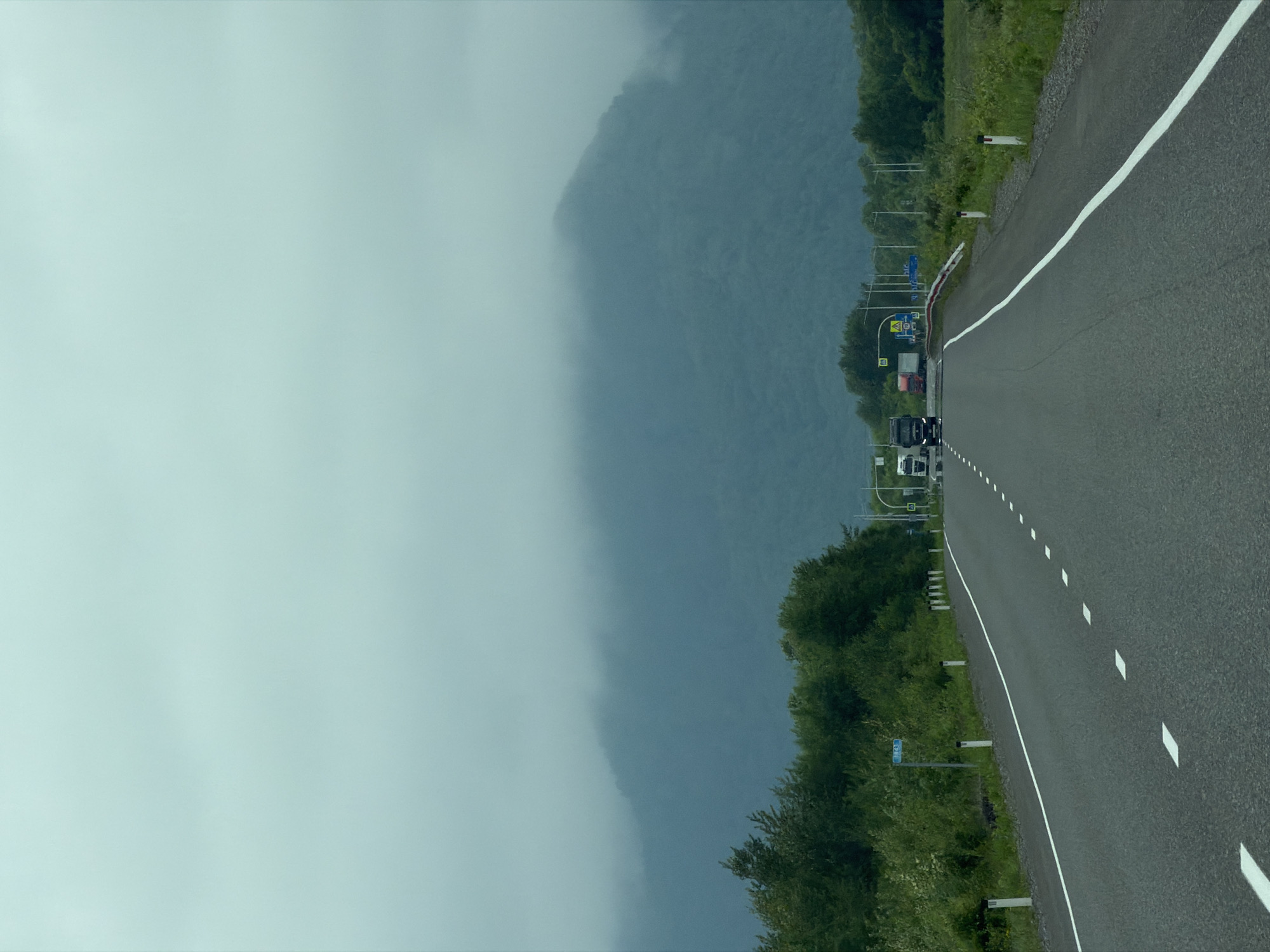 The image depicts a highway in a rural area, likely in or near Лондоко-завод, Russia. The road is paved and marked with white lines, with white post delineators along the right shoulder. On the left side of the road, there is a dense growth of green trees and shrubs, and a blue sign with white lettering is visible. Further down the road, several trucks are traveling, and there are traffic signs and lights. In the background, a large, tree-covered mountain is shrouded in mist, contributing to an overcast and atmospheric scene. The overall environment suggests a natural landscape with a functional transportation route.