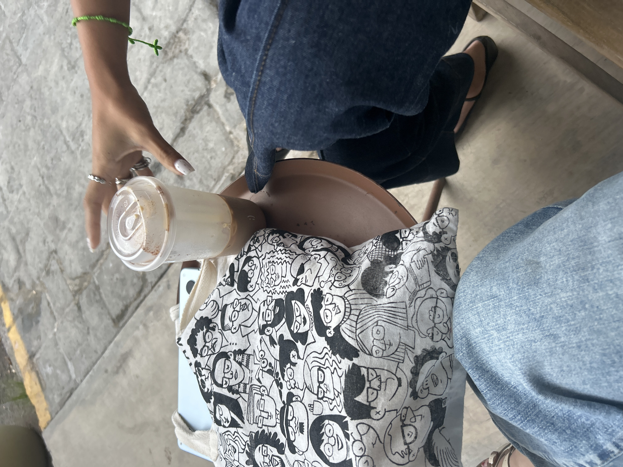 A high-angle shot depicts a person seated at an outdoor venue in Puebla, Mexico, based on the distinctive brick paving visible in the background.
The person is wearing denim jeans; one leg is dark blue and the other light blue. Footwear consists of flat sandals with tan straps. A thin, green bracelet is on the person's left wrist. Their left hand holds a clear plastic cup containing a light-colored beverage, likely coffee or a similar drink. Several rings are on their fingers.
A canvas tote bag with a black and white cartoon face design is draped over a blue object, which is positioned on the small brown table next to the person. The table is situated alongside a light-colored wooden structure.