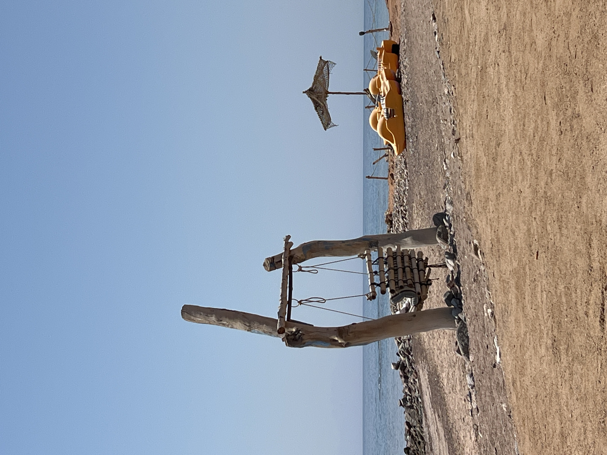 This image depicts a serene beach scene in Nuweiba, Egypt, under a clear blue sky. In the foreground, a rustic wooden swing set made of driftwood stands on a sandy and rocky shore. Hanging from the swing are several cylindrical bamboo-like objects, possibly decorative or part of a chime. To the right, in the mid-ground, a cluster of bright yellow pedal boats are lined up on the beach, partially shaded by a rustic umbrella made of netting. The ocean stretches out in the background, with gentle waves lapping against a rocky shoreline. The overall impression is one of a quiet, sun-drenched coastal retreat.