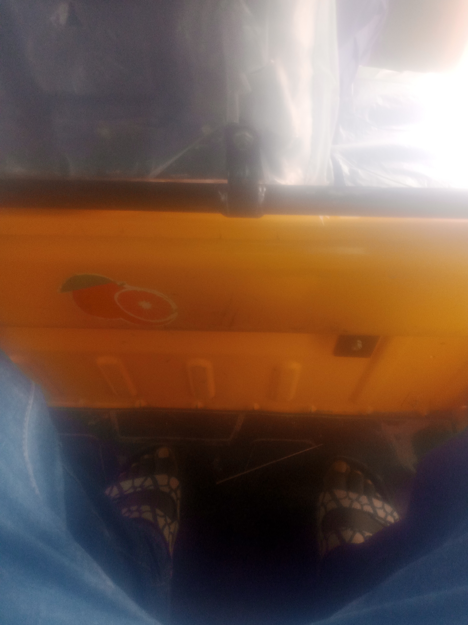 The image appears to be taken from inside a vehicle, likely a tricycle (Keke Napep) given the perspective and the bright orange panel in the foreground. The perspective is looking downwards, showing a person's feet wearing patterned sandals. One foot is visible on what appears to be a pedal or floorboard. The orange panel has a sticker depicting a stylized orange fruit with a leaf. Above the orange panel, there is a dark metal bar and then a blurry background that suggests the interior of a covered vehicle, possibly with plastic sheeting. The lighting is bright and blown out in some areas, suggesting sunlight or strong internal lighting. The image has a grainy quality. There are no identifiable landmarks or environmental cues to pinpoint a specific location within Jalingo, Nigeria. The scene is consistent with being a passenger in a Keke Napep operating within Jalingo.