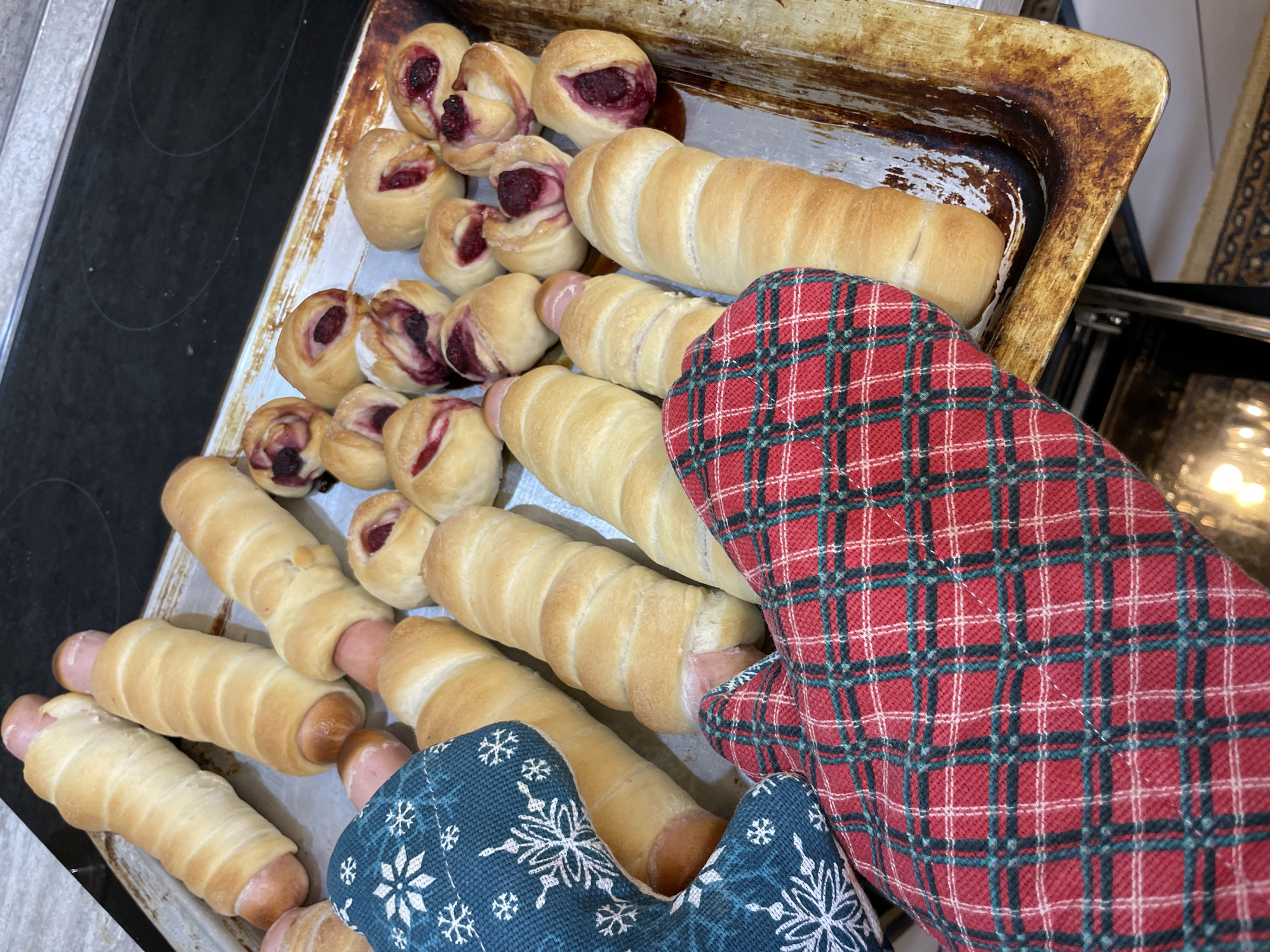 A baking sheet, held with oven mitts, contains baked goods. Some are sausage rolls, and others are round, fruit-filled pastries. The baking sheet is slightly tarnished. The oven mitts are a red plaid and a blue snowflake pattern. The scene is indoors, adjacent to a stovetop and oven. The location is not deducible as Zav'yalovo, Russia.