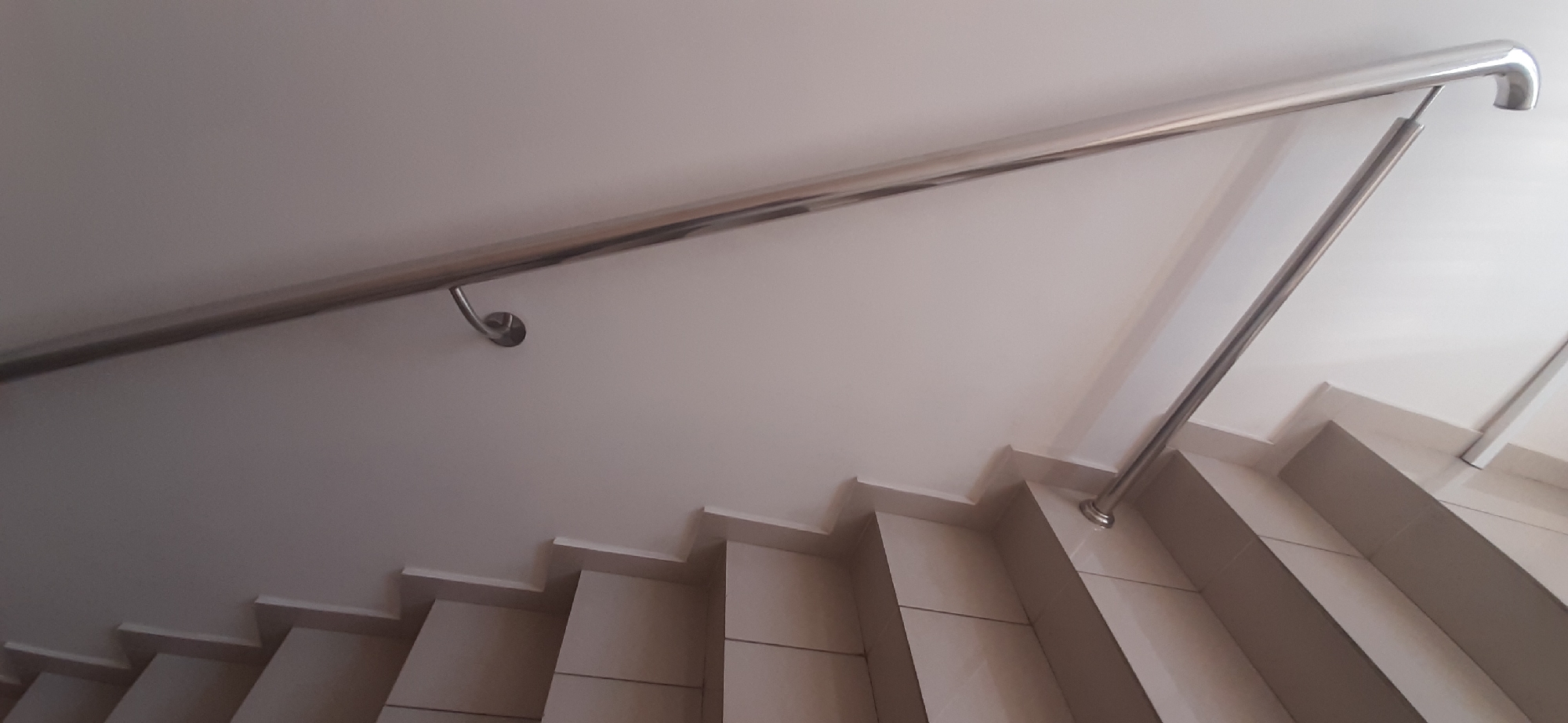 The image depicts a chrome handrail running diagonally across a white wall above a set of stairs. The stairs appear to be made of beige tiles with white risers, arranged in a zig-zag pattern. The handrail is mounted on the wall with curved brackets, and it has a rounded end on the right side of the frame. The scene appears to be an indoor staircase, possibly in a residential or public building. The lighting suggests an interior environment with ambient light. There are no people or specific markings that would indicate a precise location within Yessentuki, Russia. The image captures a common architectural feature.
