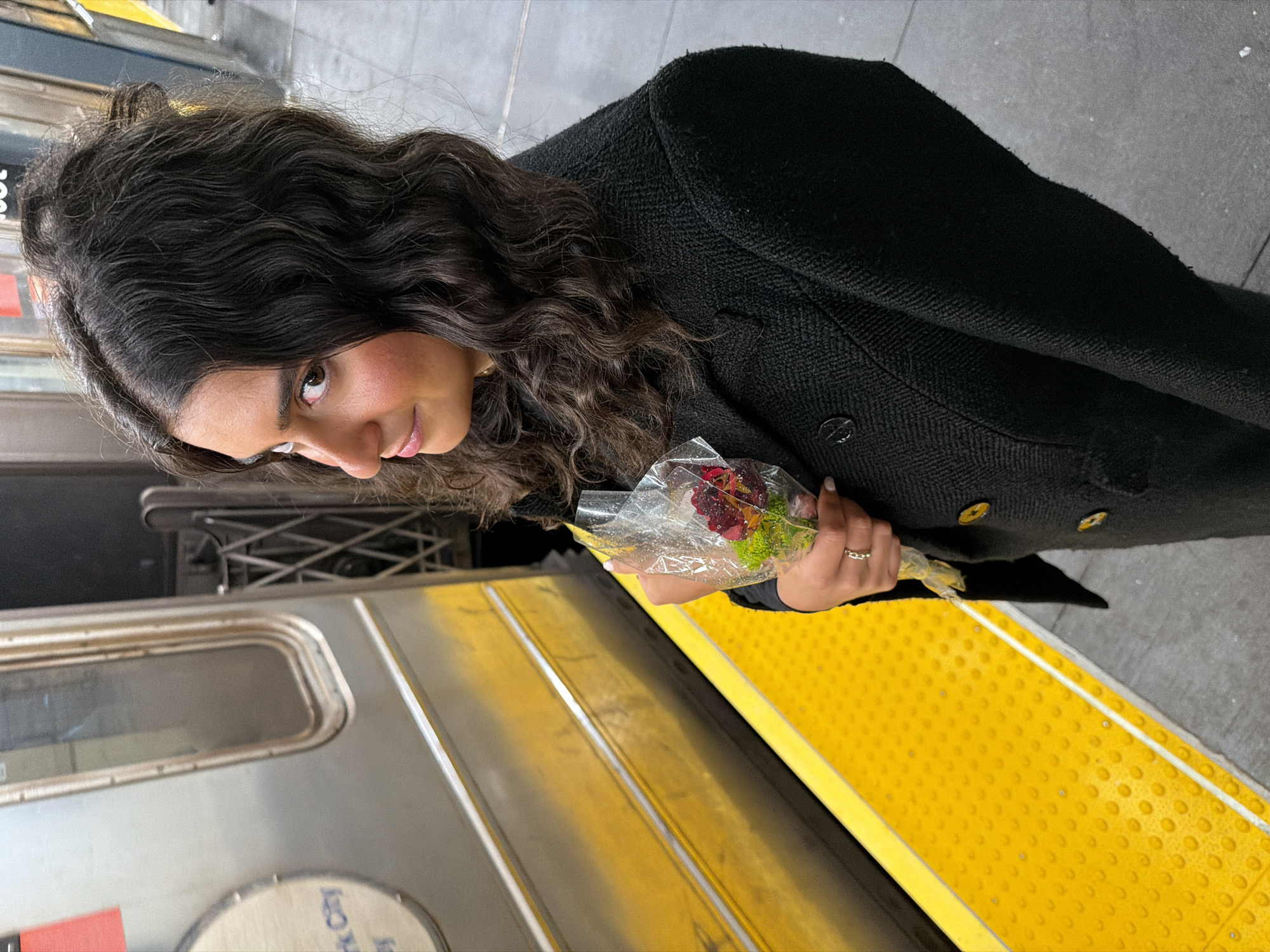 A woman with long, dark, curly hair is standing on a New York City subway platform, holding a small bouquet of flowers wrapped in clear plastic. She wears a black coat with gold buttons. The subway train is visible to the left, with the yellow safety strip on the platform in the foreground. The platform surface is grey.