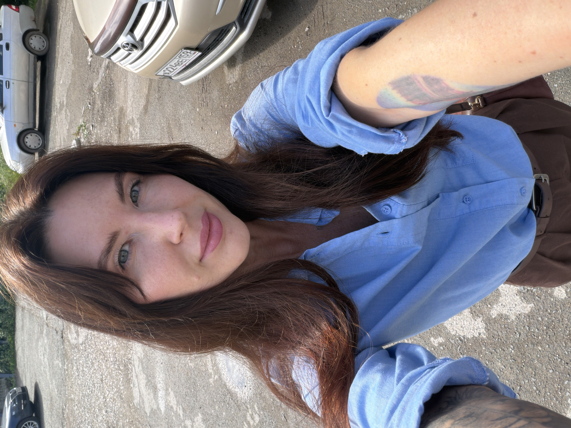 A woman is taking a selfie outdoors in what appears to be a parking lot. She has long, dark brown hair and is wearing a light blue button-down shirt and brown pants. A portion of a beige car is visible in the background, along with a silver car parked further away. The ground is asphalt with some gravel and scattered debris. There is a blurry tattoo on her right arm. The license plate of the beige car reads "A224BE159", suggesting the location could be Perm, Russia, as the "159" region code corresponds to Perm Krai. The lighting suggests it is daytime and sunny.