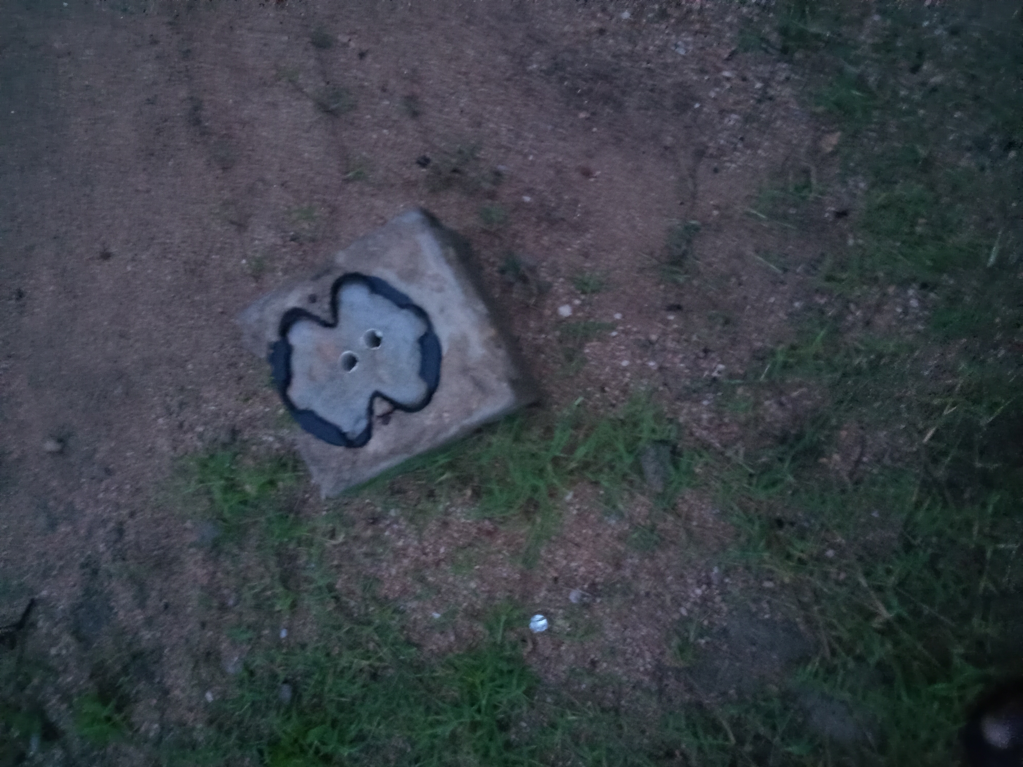 The image shows a concrete block with a circular indentation on top. The indentation contains a darker material, possibly asphalt or tar, with two visible holes in its center. The block is situated on the ground, which is a mixture of sand, gravel, and sparse green grass. The overall lighting suggests it is either early morning or late evening. The scene appears to be outdoors, possibly in a semi-urban or rural setting within Jalingo, Nigeria, given the information provided. There are no people or animals visible in the frame. The composition is a top-down view of the concrete block and its immediate surroundings.