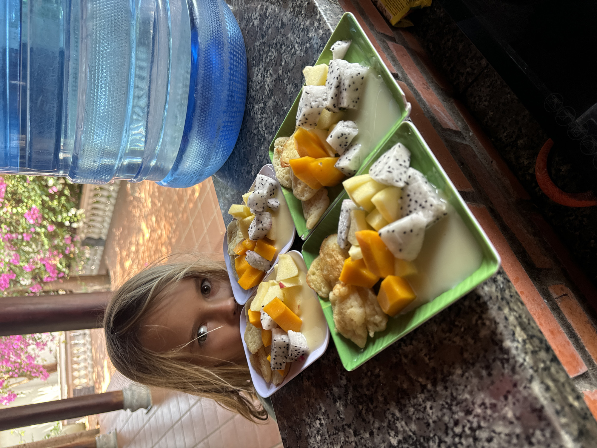 The image, taken in Mũi Né, Vietnam, is rotated 90 degrees clockwise and captures a close-up scene featuring a young person and several bowls of fruit. A child with light-colored hair and wide, curious eyes is visible on the left side of the frame, looking directly at the camera.
On a dark, speckled granite-like countertop in the midground, there are multiple servings of fruit salad. These salads contain chunks of white dragon fruit with black seeds, diced yellow mango, and other light yellow fruit, all immersed in a light, creamy liquid (possibly yogurt or coconut milk). Flat, round, biscuit-like items are also present in the bowls. Two of the servings are in square, bright green dishes, while two others are in smaller, white oval dishes.
On the far left of the counter, a large, blue plastic water cooler bottle is prominently featured. The background on the left, behind the child, shows lush green foliage with vibrant pink flowers, suggesting an outdoor or semi-outdoor setting, possibly a patio or balcony, with a dark railing also visible. To the right, behind the fruit bowls, a reddish brick wall can be seen. The bright, clear lighting indicates that the scene is taking place during the daytime. No legible text is visible in the image.