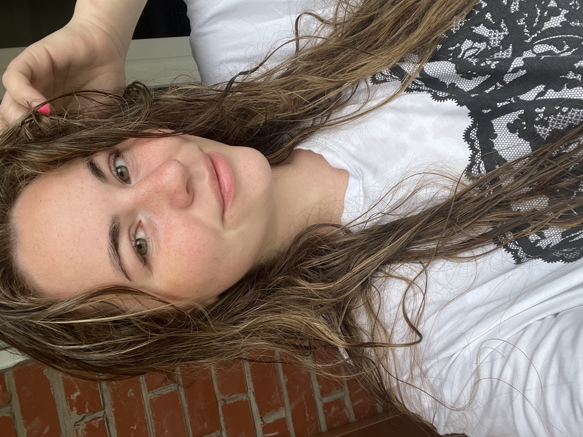 A young woman with damp, light brown hair and green eyes smiles at the camera. She has freckles across her nose and cheeks and is wearing a white t-shirt with a black lace design on the front. Her left hand is raised, touching her hair. She is standing in front of a red brick wall. The background suggests an outdoor urban setting, possibly a residential area in Chaikovsky, Russia, given the typical architecture and brickwork found in the city.