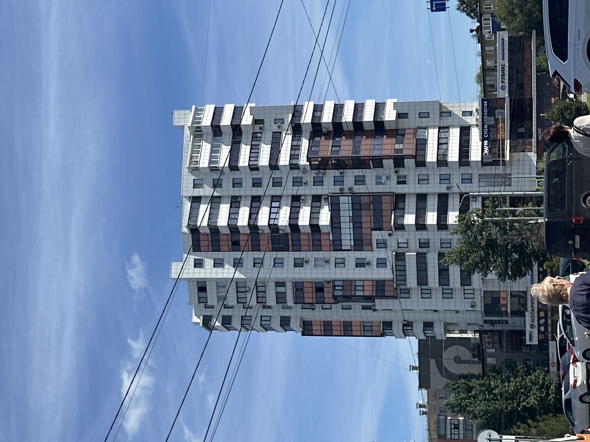 An eye-level, medium shot captures a tall, modern apartment building in an urban environment under a bright blue sky with scattered clouds. The building, characterized by its white exterior panels and numerous balconies with tinted windows, dominates the center of the frame. Electrical wires crisscross the foreground and extend across the building's facade. To the left of the apartment building, a lower, older building with a large circular graphic is visible, partially obscured by green trees. In the foreground, cars are parked and driving on a street, and two individuals are walking away from the camera, with their backs to the viewer. The scene suggests a typical city street during the daytime.