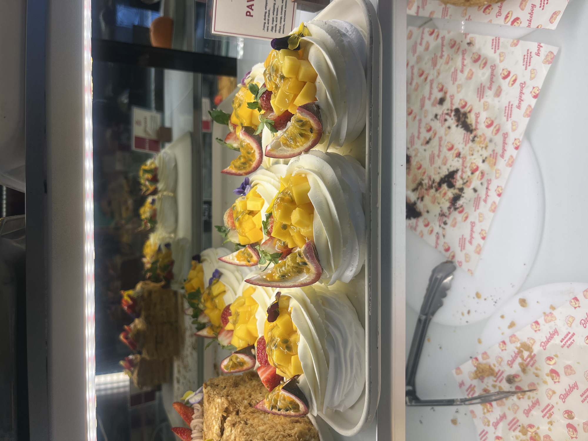 A close-up shot captures a display of desserts inside a glass case, likely located in a shop called "Bakeboy." The desserts are primarily pavlovas, a meringue-based dessert, adorned with fresh fruit, including mango chunks, strawberries, and passion fruit slices. The pavlovas are arranged in a row and sit atop what appears to be another layer of pastry. A sign with the label "PAV" can be seen, likely referring to "Pavlova," with ingredients listed in multiple languages. The background reveals more pastries and cakes, further indicating a bakery or patisserie setting. Tongs are visible in the case, suggesting customer service. The branding of "Bakeboy" is repeated on the napkins, reinforcing the location.