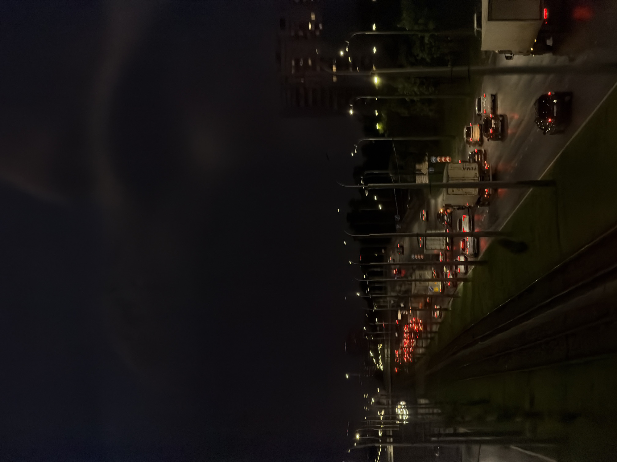 An overhead shot captures a nighttime scene of a highway in an urban environment. The highway is filled with vehicles, their headlights and taillights creating streaks of light against the dark asphalt, indicating slow-moving or stopped traffic. Streetlights line the highway, illuminating the scene with a warm, yellow glow. In the background, a tall building with illuminated windows stands against the dark sky. The overall impression is one of a busy city at night, with the glow of artificial lights contrasting with the darkness of the night sky.