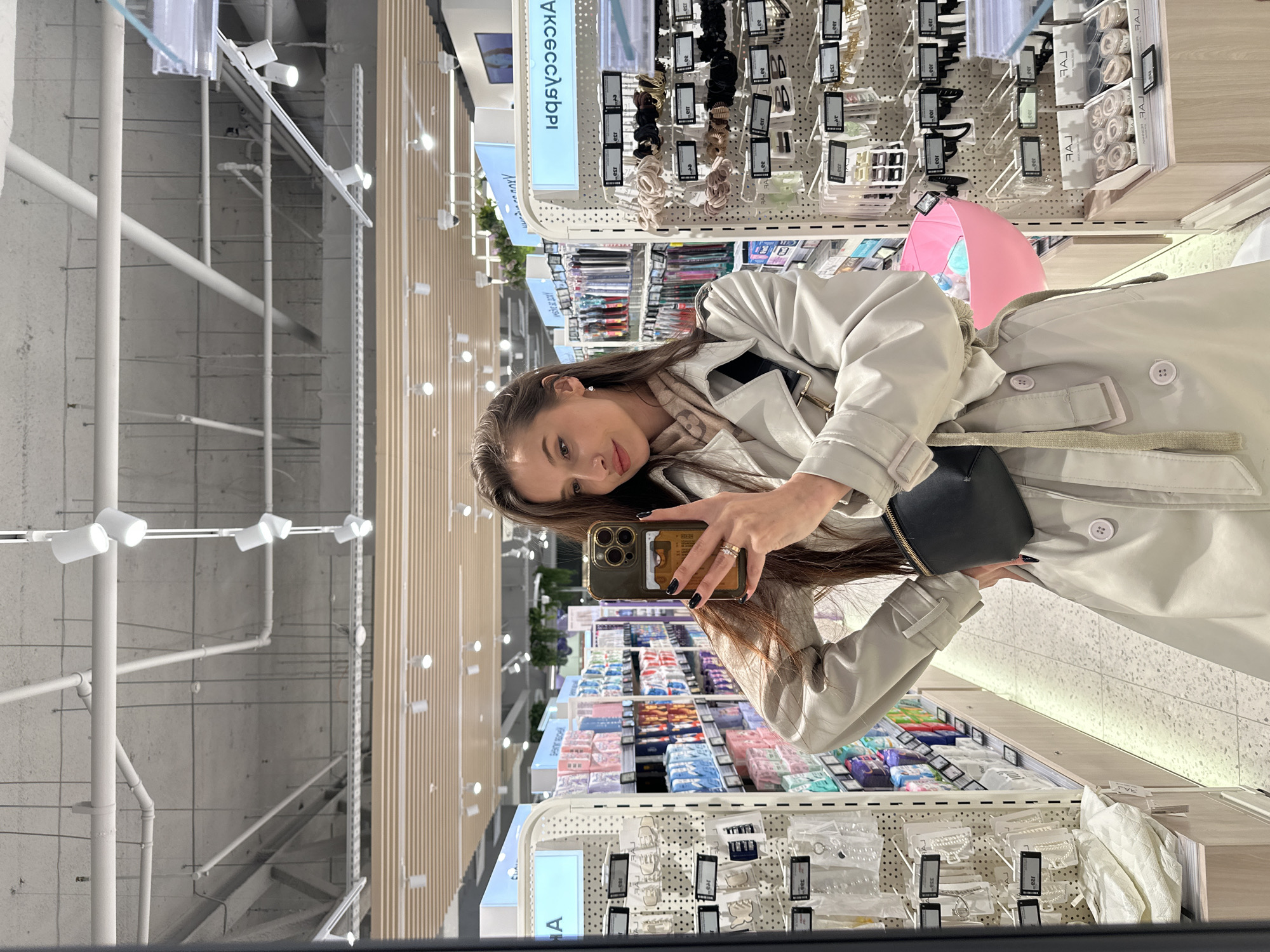 A young woman is captured in a selfie taken in what appears to be a mirror or reflective surface within a retail store. The image is oriented sideways, with the woman looking directly at the camera, holding a smartphone with a prominent camera array and dark phone case. Her dark hair falls over her shoulders, and she wears dark nail polish, a ring on her right hand, and a light-colored, possibly beige or off-white, trench coat over a patterned scarf. A small black shoulder bag is visible on her side.
The setting is a well-lit interior of a store, possibly a beauty or accessories section. Behind the woman, numerous display racks hold various items, predominantly hair accessories like scrunchies, clips, and other small ornaments, packaged individually. The merchandise appears to be neatly organized on white grid-like displays. One large sign in the background clearly reads "АКСЕССУАРЫ," which is Russian for "ACCESSORIES," indicating the section. Smaller labels on products also show fragmented text like "ТЭГ" (TAG) and "ЛАЙК" (LIKE).
Further in the background, shelves are stocked with colorful packaged goods, likely personal care items or similar small merchandise. Above, the store's ceiling features exposed white pipes and track lighting, creating a modern industrial aesthetic. To the right of the woman, a partial view of a bright pink umbrella adds a pop of color to the otherwise neutral store environment. The overall impression is a casual moment during a shopping trip in a modern retail space.