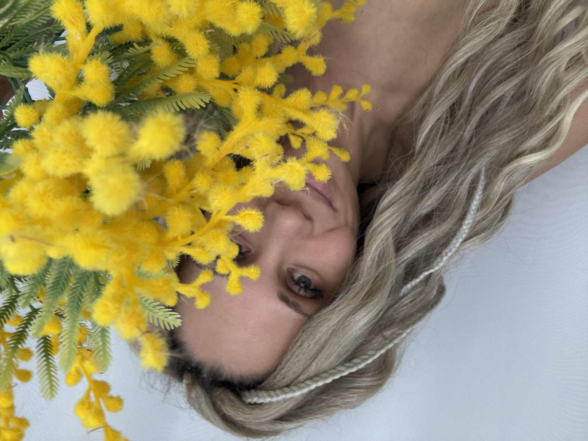 A medium-shot portrait reveals a woman with fair skin, adorned with blonde dreadlocks accentuated with braided strands of white thread. Her gaze is directed towards the camera, with her partially obscured facial features including her eyes and the left side of her mouth. The woman is positioned against a backdrop of yellow Mimosa flowers, their green foliage intermingling with the vibrant yellow blooms. The scene gives a sense of intimacy. The location is not mentioned in the photo and is not determinable.
