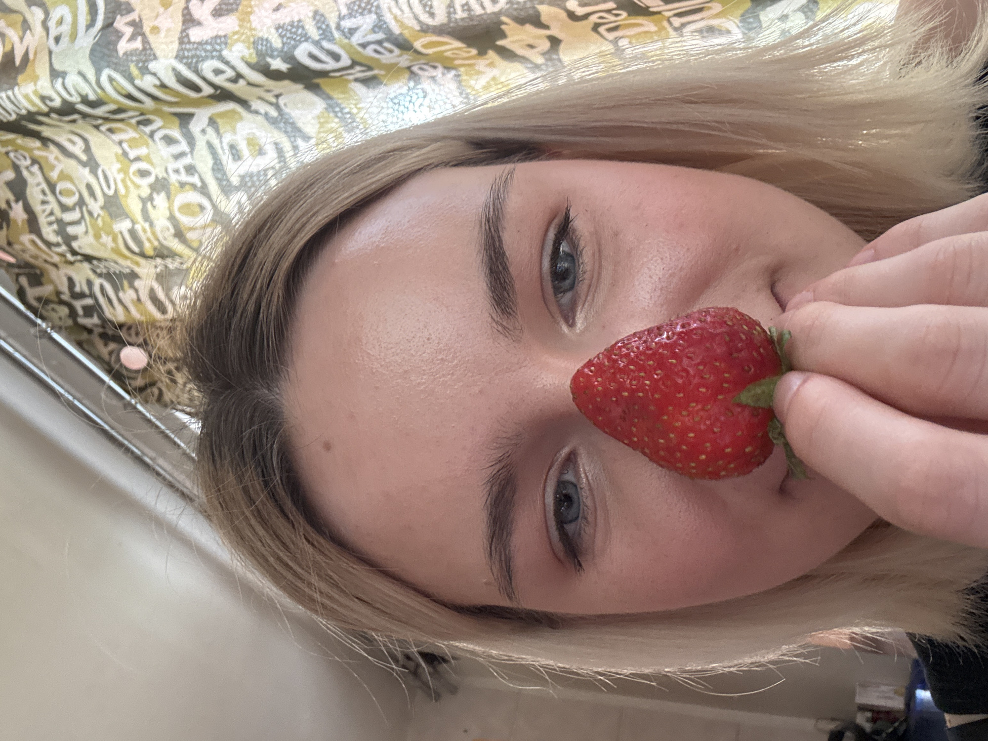 The image presents a close-up, tilted portrait of a person, likely a young woman, with blonde hair and light blue-grey eyes. The subject is holding a bright red strawberry with their right hand, positioning it playfully against their nose and upper lip. Their expression is neutral to slightly smiling, conveying a casual and perhaps whimsical mood.
The background consists of a patterned fabric or curtain with abstractly arranged and repeated text in yellow, green, and cream hues. Partially visible words and fragments include "ORDER," "wonder," "well," "of," "adorn," and "the." To the far left, a light-colored surface, possibly a wall, and narrow metallic bars (suggesting a railing or fixture) are visible.
The lighting is bright and even, indicating an indoor setting, likely during daytime. The subject's blonde hair is casually styled around their face. The overall aesthetic is informal and intimate. While the provided context notes the location as Saint Petersburg, Russia, there are no specific visual cues within the image itself to confirm this.