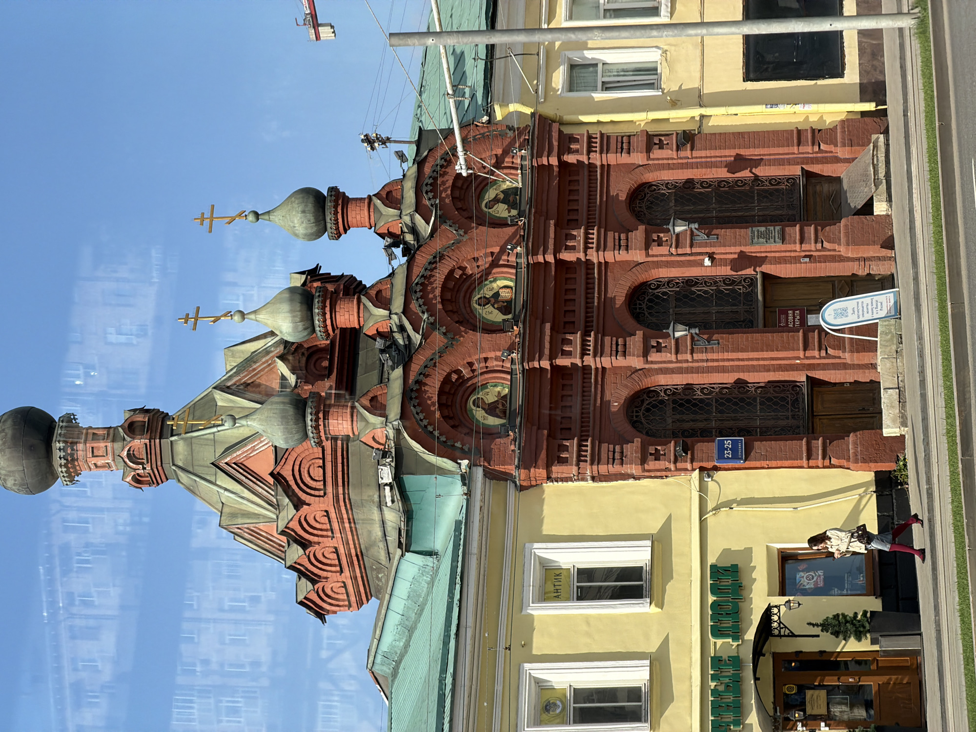 A vertical eye-level shot shows a church and adjacent building in Moscow, Russia, likely taken through a window. The sky is visible on the left, reflecting off of the window and appearing light blue. The church is a red brick building with green roofs on some of its sections and domes topped with crosses. The adjacent building is painted yellow. On the building, a sign with Cyrillic letters hangs above a doorway that reads "АНТИК." A woman in red tights walks past the storefront, holding a cell phone and wearing a light-colored jacket. A small street sign is visible next to a closed doorway that reads "23-25." The image appears to have been taken in daylight, but the exact time is unknown.