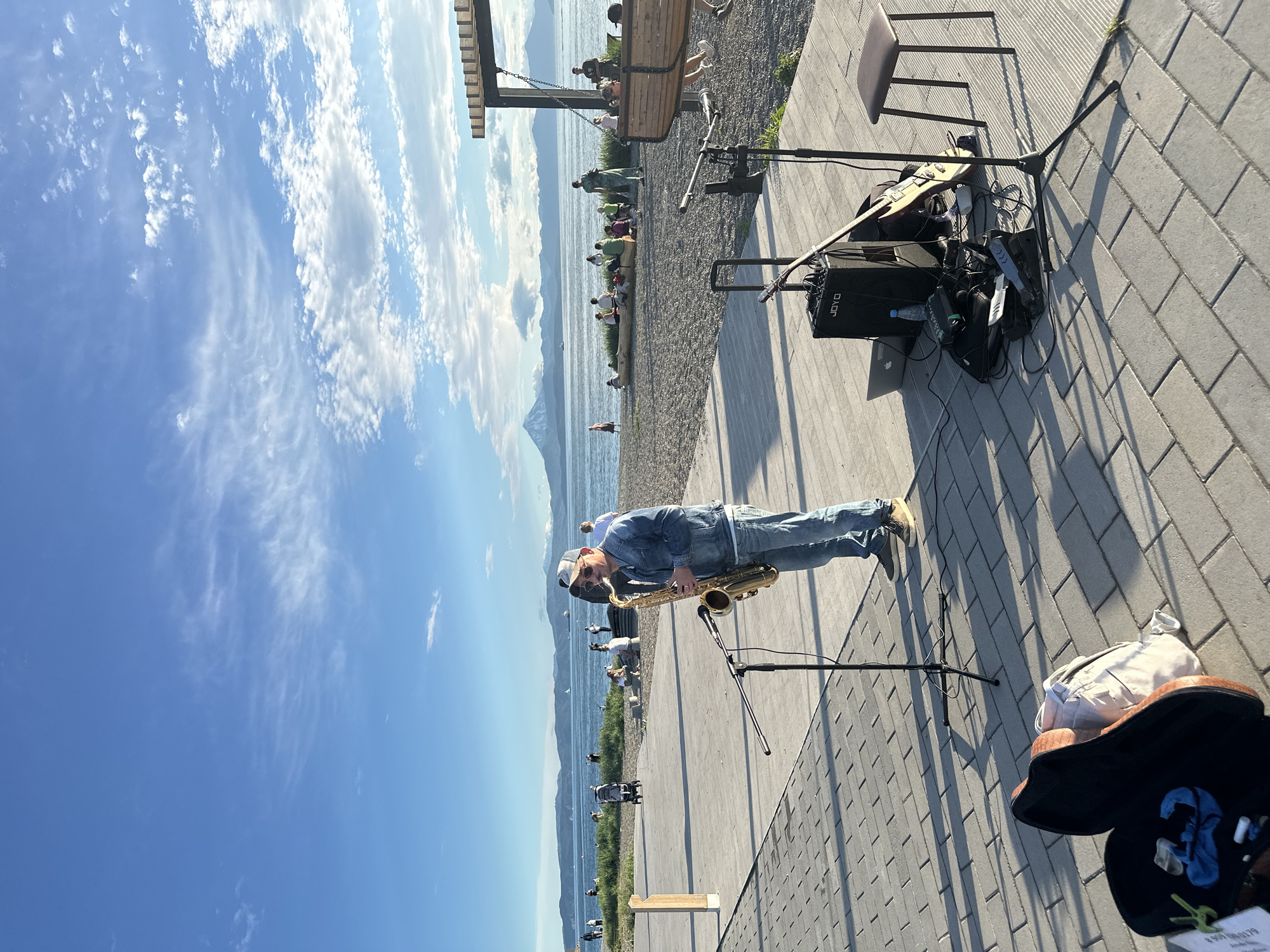 A male saxophonist stands on a paved walkway, playing his instrument. He is wearing a denim jacket, jeans, a baseball cap, and sunglasses. In front of him are microphones, a portable amplifier, a laptop, an electric guitar, and other musical equipment. A guitar case and a backpack are on the ground to his left. The walkway overlooks a beach area with people sitting and standing, some gathered around a large piece of driftwood. In the background, the ocean stretches to the horizon, with mountains and a prominent volcano visible under a partly cloudy blue sky. The scene appears to be in Petropavlovsk-Kamchatsky, Russia, likely along the coast of Avacha Bay.