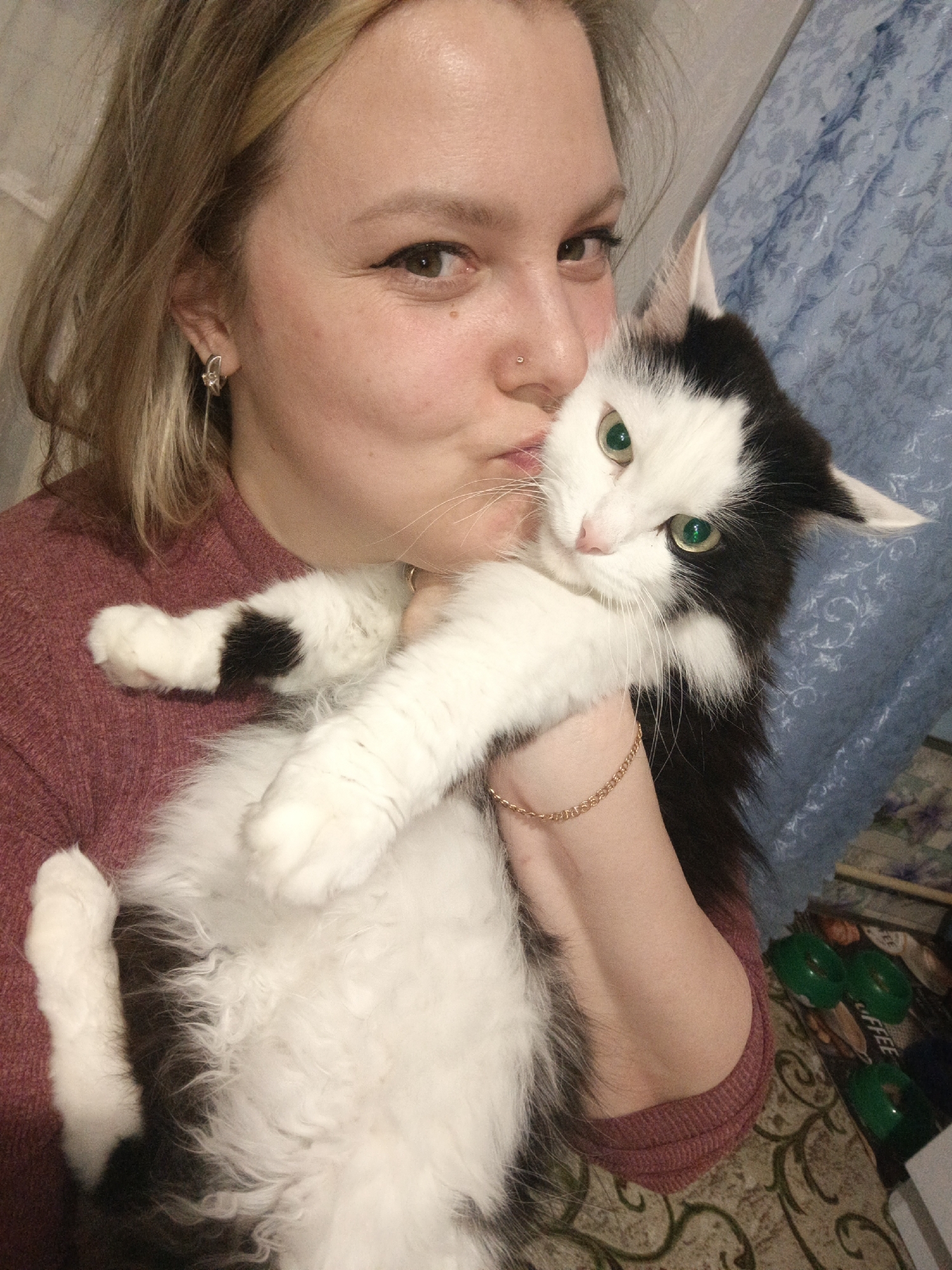 An affectionate moment is captured indoors, likely in a home in Markovskiy, Russia, featuring a woman holding a cat. The woman, with blonde hair and fair skin, wears a reddish-purple ribbed sweater. She has a subtle nose piercing, an earring in her left ear, and a thin gold chain bracelet on her right wrist. Her eyes are open, looking towards the camera, while her lips are puckered as if she is kissing or nuzzling the cat's head.
She cradles a fluffy, long-haired black and white cat. The cat has striking bright green eyes and prominent white whiskers. Its face, chest, and belly are predominantly white, while its head, ears, and back are black. Some black patches are visible on its white paws. The cat appears to be looking directly at the camera with wide eyes, its ears slightly back, suggesting a moment of mild surprise or tolerance rather than distress.
The background reveals a domestic setting. Behind the subjects, a blue patterned fabric, possibly a curtain or blanket, is visible. To the right, a patterned carpet or rug with green and brown motifs covers the floor. On the floor, some objects are discernible, including green plastic lids and packaging that clearly displays the word "COFFEE". The lighting suggests an indoor setting, likely under artificial illumination. The overall scene conveys a warm, personal interaction between a pet owner and her cat.