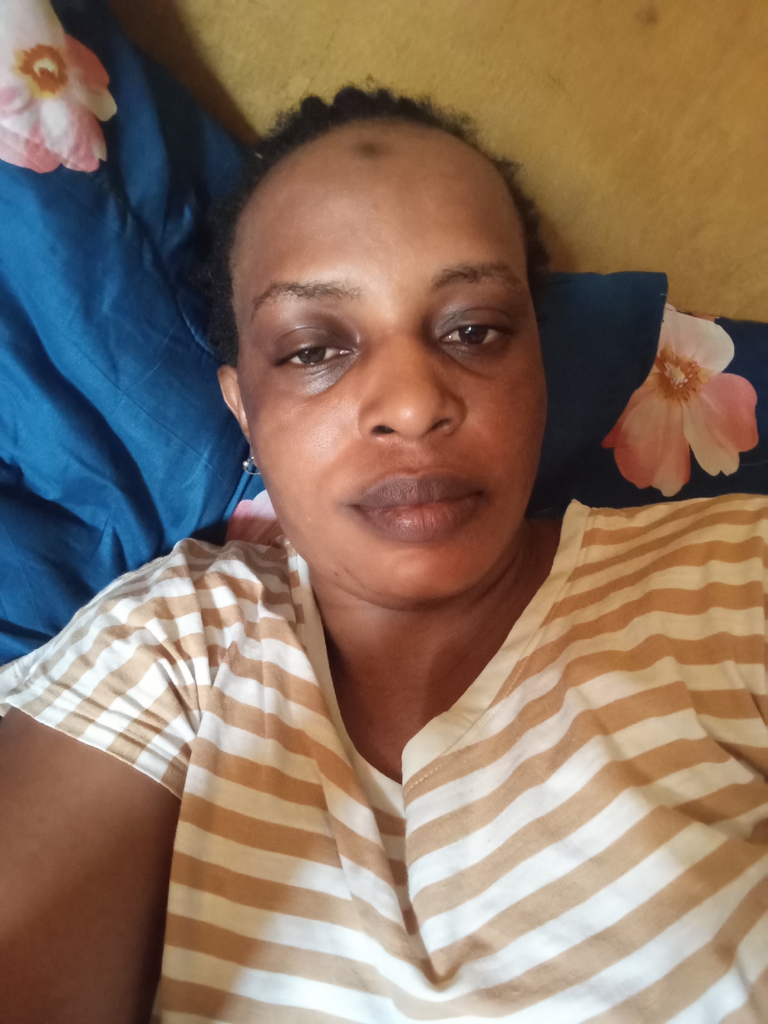 A close-up, eye-level selfie captures a woman with dark skin and dark hair. She is wearing a striped, short-sleeved shirt. She is lying on a bed, her head on a blue pillow with a floral pattern. The background includes a yellow wall. The location is Narayi, Nigeria.