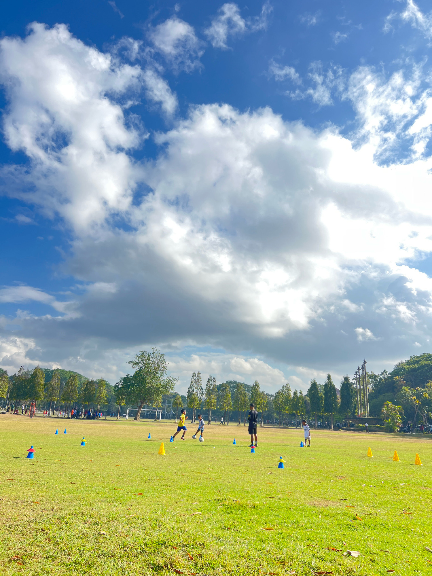 This image depicts a soccer practice taking place on a grassy field under a partly cloudy blue sky. In the foreground, several colorful cones are scattered across the field, indicating a training drill. Two young boys are actively engaged in playing soccer, with one dribbling a ball while the other runs towards it. Further in the background, other individuals can be seen on the field, some appearing to be coaches or participants in further drills. A soccer goal is visible in the distance. The background is framed by a line of trees and lush green foliage, suggesting a park or sports facility. The presence of a statue-like monument in the far background, along with the style of architecture in the distance, suggests this location could be the Puputan Badung Monument Park in Denpasar, Bali, Indonesia, a common public space for recreational activities. The overall scene is one of energetic activity and outdoor recreation in a well-maintained park setting.