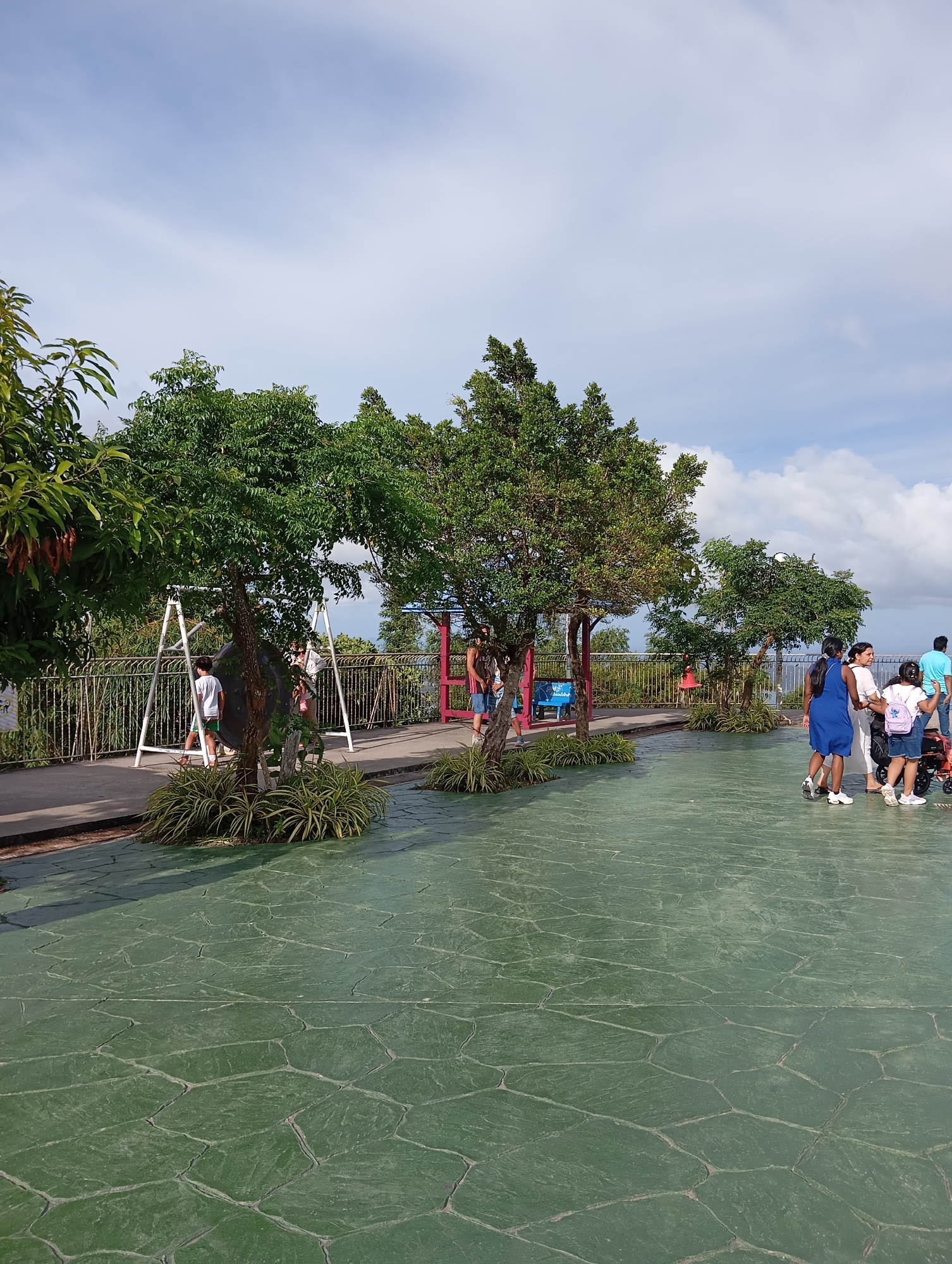 This image depicts a bright, sunny day at a scenic overlook in Karon, Thailand, with a clear blue sky dotted with white clouds. In the foreground, a textured green concrete patio with a stone-like pattern dominates the lower half of the frame. A group of people are walking on the patio towards the right side of the image. One woman in a blue dress is seen from the side, holding the hand of a young girl wearing a backpack. Next to them, a man in a light blue shirt walks away from the camera, and a woman in a white shirt is also present. Further left, a man in shorts and a dark t-shirt stands near a stroller.
Beyond the patio, a paved walkway runs horizontally across the mid-ground, bordered by a metal fence. Several trees with lush green foliage are strategically placed along the walkway and on small mounds of ornamental grass. To the left, a white swing set with a large black sphere is visible, with two children standing near it. In the background, a vibrant pink structure stands out, and a hazy landscape suggests a distant view, possibly of the sea. The overall atmosphere is one of leisure and outdoor enjoyment.