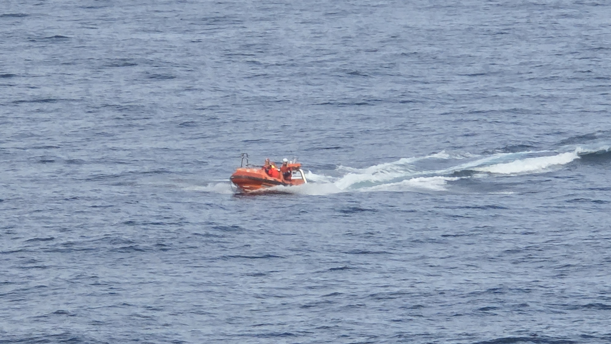 A photograph shows an orange inflatable rescue boat speeding across a body of water. The boat is surrounded by white foam and water spray generated by its movement. There appear to be at least three people on board, all wearing what look like personal flotation devices. The surrounding water is a deep blue, with small waves visible across the surface. The location is Unknown, Unknown.