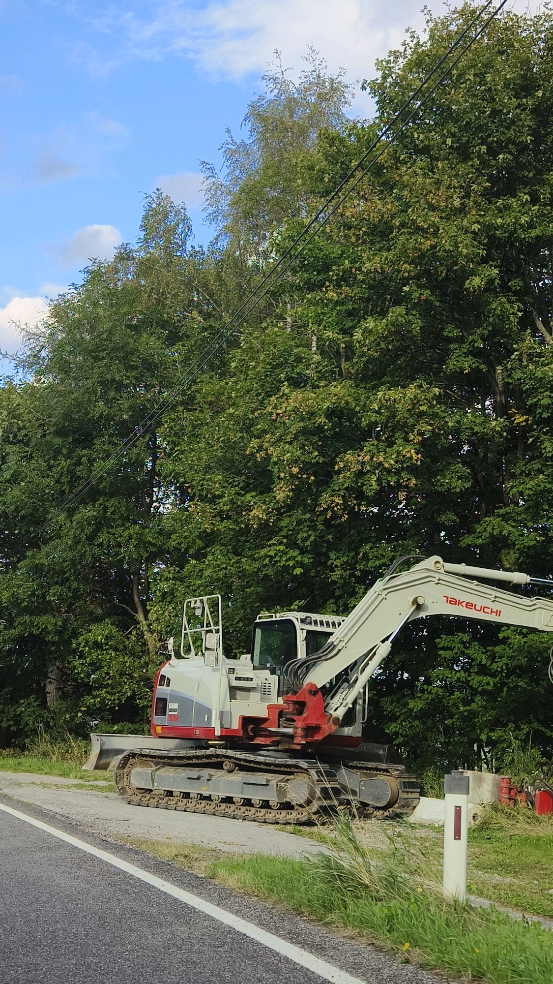 An excavator is parked on the side of a paved road bordered by grass and dense trees under a partly cloudy sky. The excavator is white and red with the brand name "TAKEUCHI" visible on its arm. A white post with a red marker is situated in the grass to the right of the excavator. The scene appears to be located near Schottwien, Austria, as suggested by the surrounding environment and the presence of construction equipment.