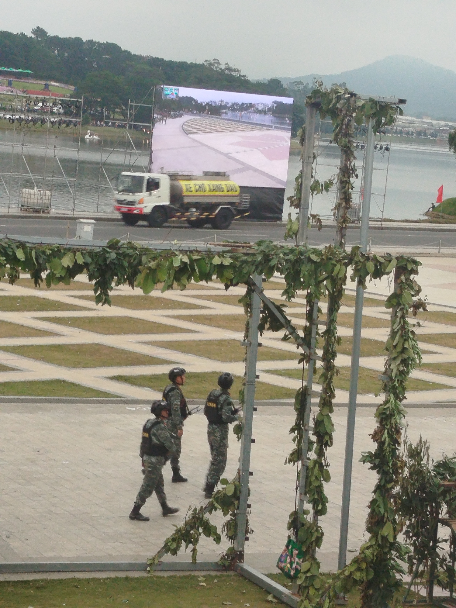 The image depicts an outdoor scene in Da Lat, Vietnam, with a large LED screen displaying a race track. In the foreground, three individuals in tactical gear and helmets are walking on a paved area. They are wearing camouflage uniforms and vests. To the left, a white truck with a yellow tank is parked. Behind the truck, a body of water is visible with a flock of swan-shaped paddle boats on it. In the background, hills covered in trees rise against a hazy sky. A red flag is planted on a grassy embankment to the right. The overall atmosphere suggests a public event or gathering, possibly a celebration or sporting event, taking place in Da Lat.