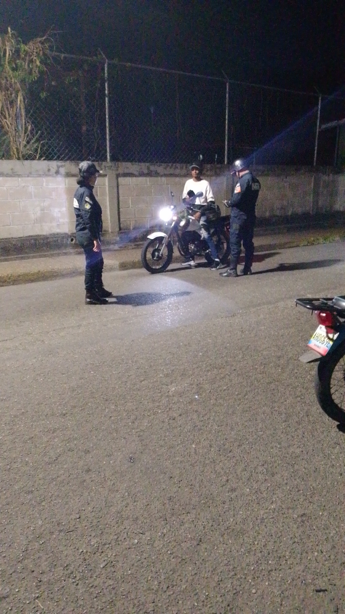 The image depicts a nighttime scene on what appears to be a paved road or street. Three individuals are visible: two police officers and one civilian. On the left, a police officer in a dark uniform, cap, and boots stands facing towards the center. In the middle, a civilian wearing a white long-sleeved shirt and a cap is seated on a white and black motorcycle. The motorcycle's headlight is brightly illuminated, casting a strong beam onto the asphalt directly in front of it. On the right, a second police officer, dressed in a dark uniform and a helmet, stands facing the civilian, seemingly engaged in an interaction.
In the background, a low concrete block wall is topped with a chain-link fence, which stretches across the scene into the darkness. Some sparse vegetation is visible behind the fence on the left side. The ground is dark, coarse asphalt. A second motorcycle is partially visible in the bottom right corner of the frame, showing its rear wheel and a license plate.
The activity taking place appears to be a police stop or official interaction with a motorcyclist. The bright headlight of the motorcycle is a dominant feature, indicating the dark conditions. The visible text on the license plate of the partial motorcycle in the foreground reads "AH0684E". The overall setting suggests an outdoor, roadside environment at night, in Tinaquillo, Venezuela, as per the provided context.