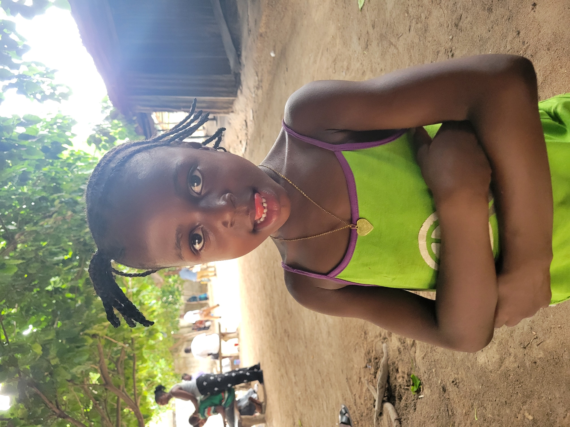 A young African girl stands outdoors on a dusty, sandy terrain, her arms crossed. She is wearing a lime green tank top with purple straps and a gold necklace with a heart-shaped pendant. Her hair is styled in two braided pigtails, and she is sticking her tongue out slightly. In the background, people are seated on chairs in what appears to be a gathering or event. A wooden structure is partially visible behind the girl, and trees with dense foliage are also present in the background. This image was taken in Madalla, Nigeria.